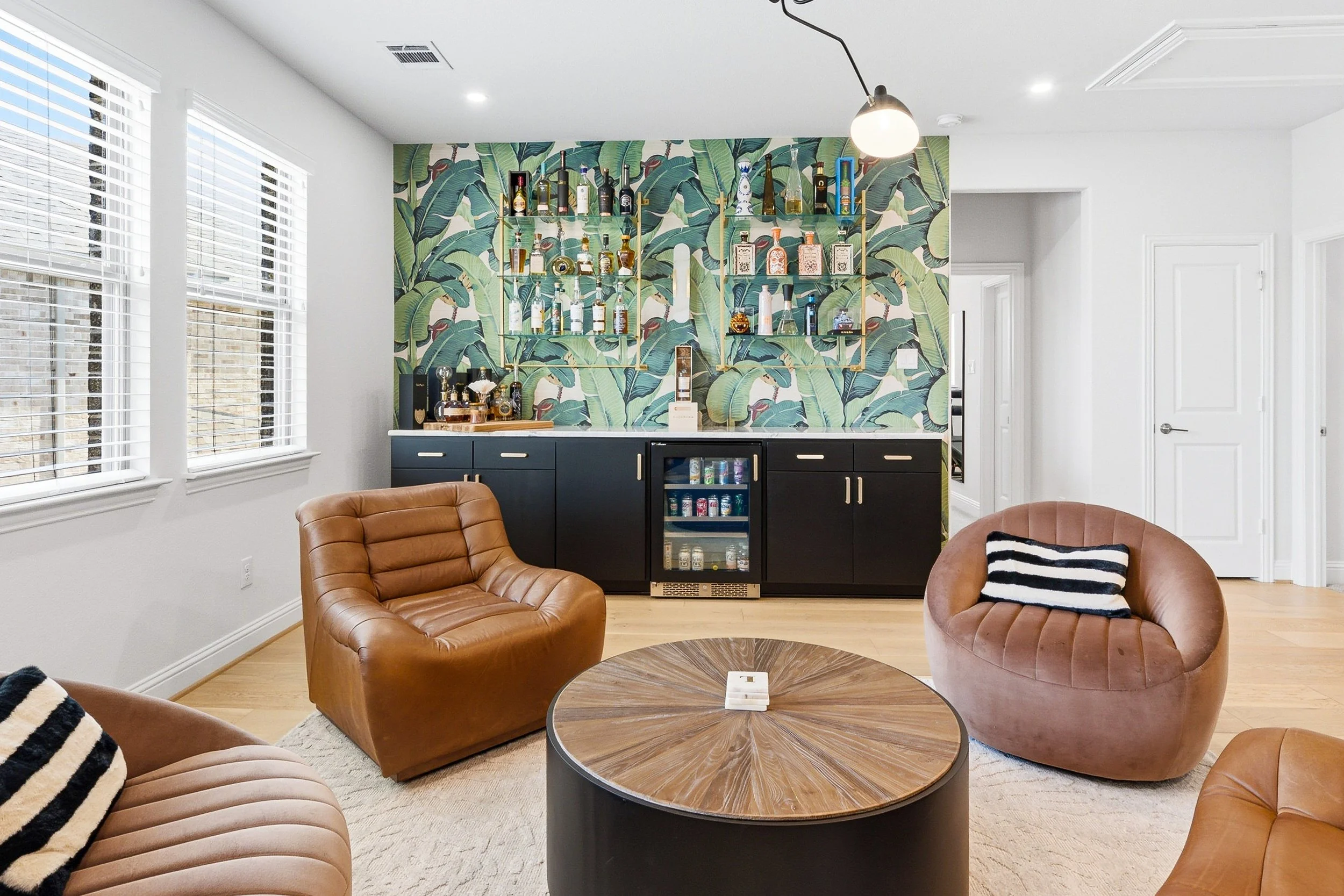 Living room with banana leaf wallpaper, black bar cabinet with drink bottles, brown leather armchairs, round wooden coffee table, large windows with white blinds, white walls and ceiling, modern light fixture.