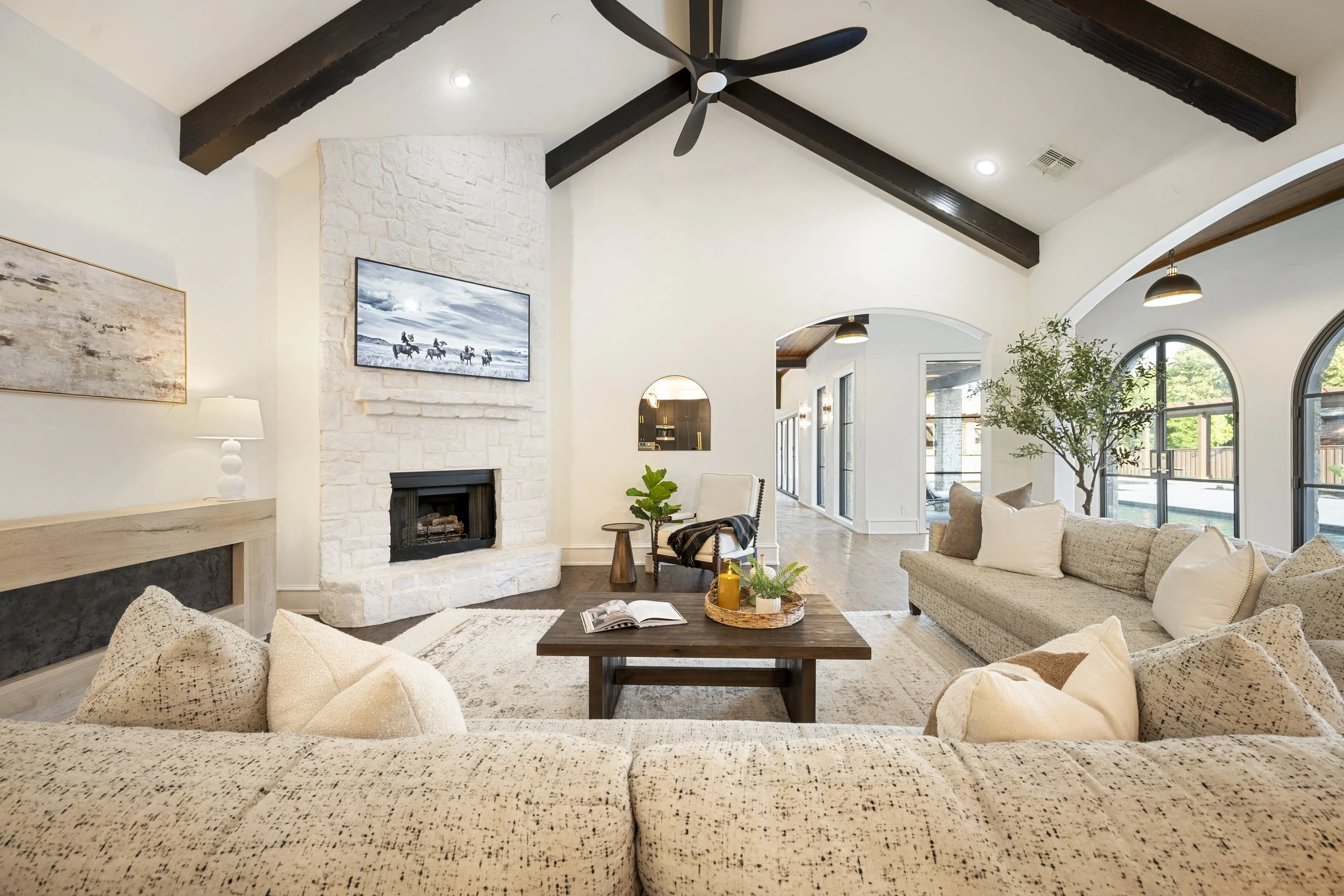 Interior view of a spacious living room with high vaulted ceiling, dark wooden beams, a black ceiling fan, white painted stone fireplace, large arched windows, and neutral-colored furniture and decor.