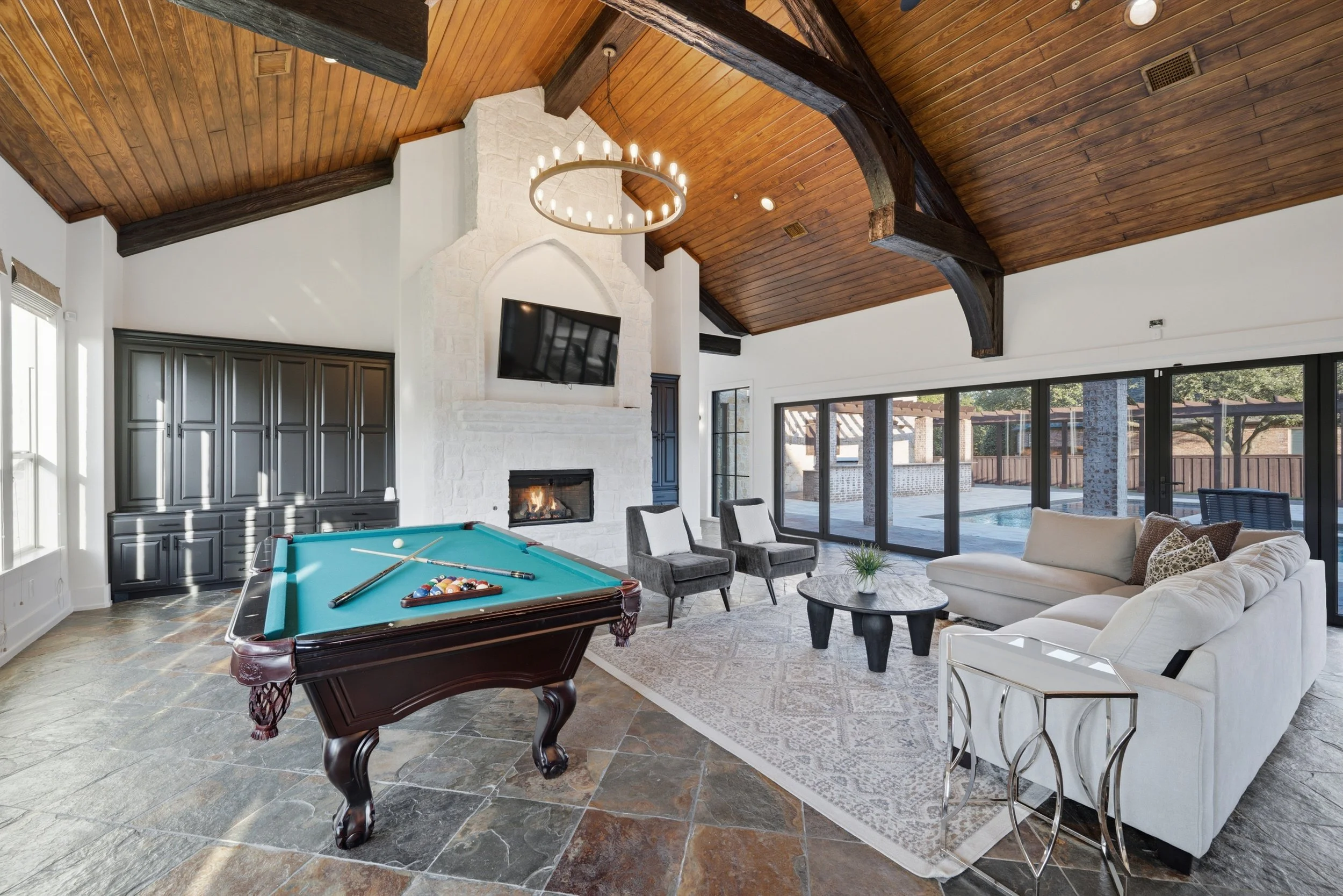 Living room with a high wooden ceiling featuring dark wooden beams, a white stone fireplace, a wall-mounted flat-screen TV, large windows with black frames, a pool table with pool balls and cues, gray chairs, a white sofa with decorative pillows, a r