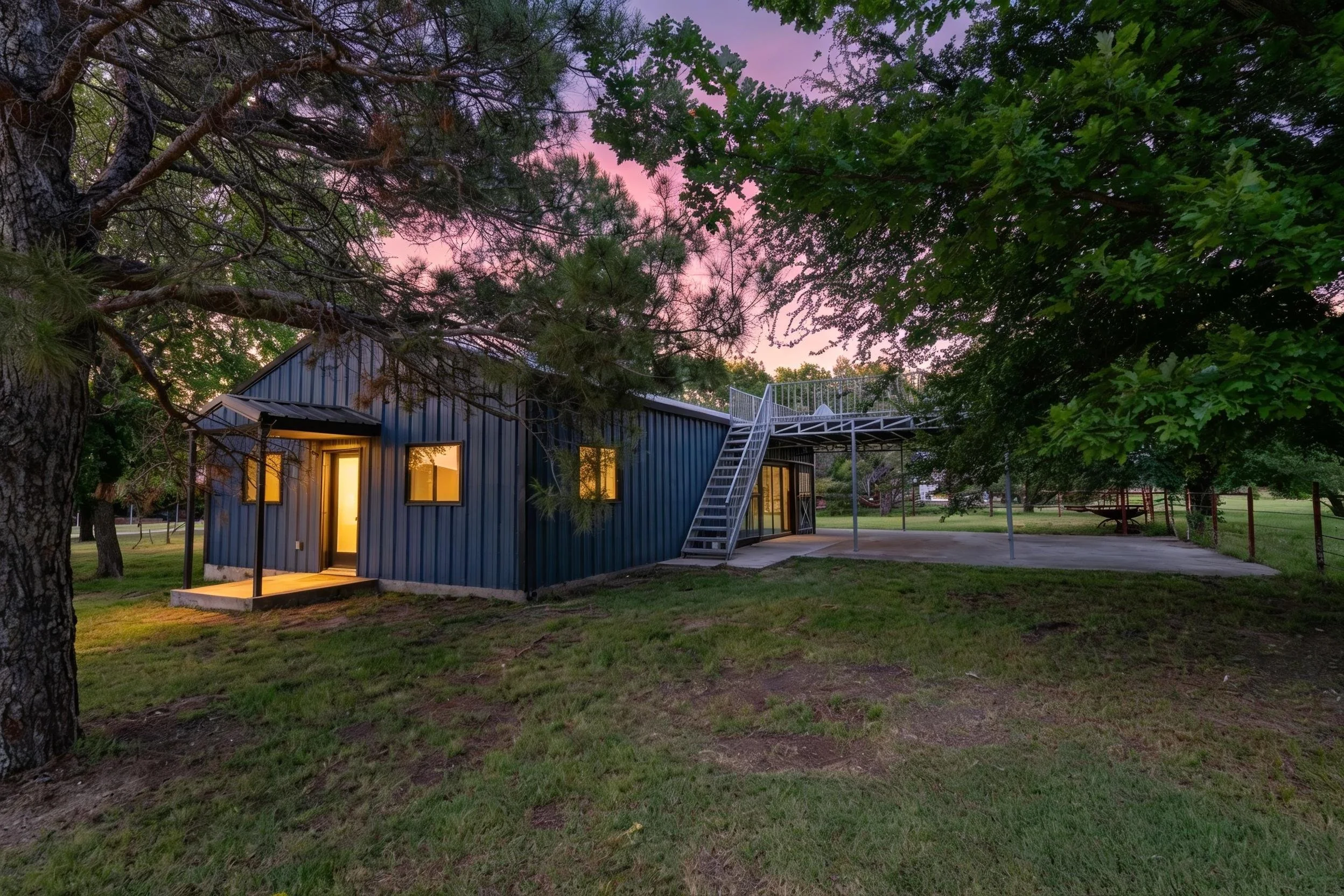 A modern blue metal building with large windows and a staircase, surrounded by trees at dusk with a purple sky in the background.