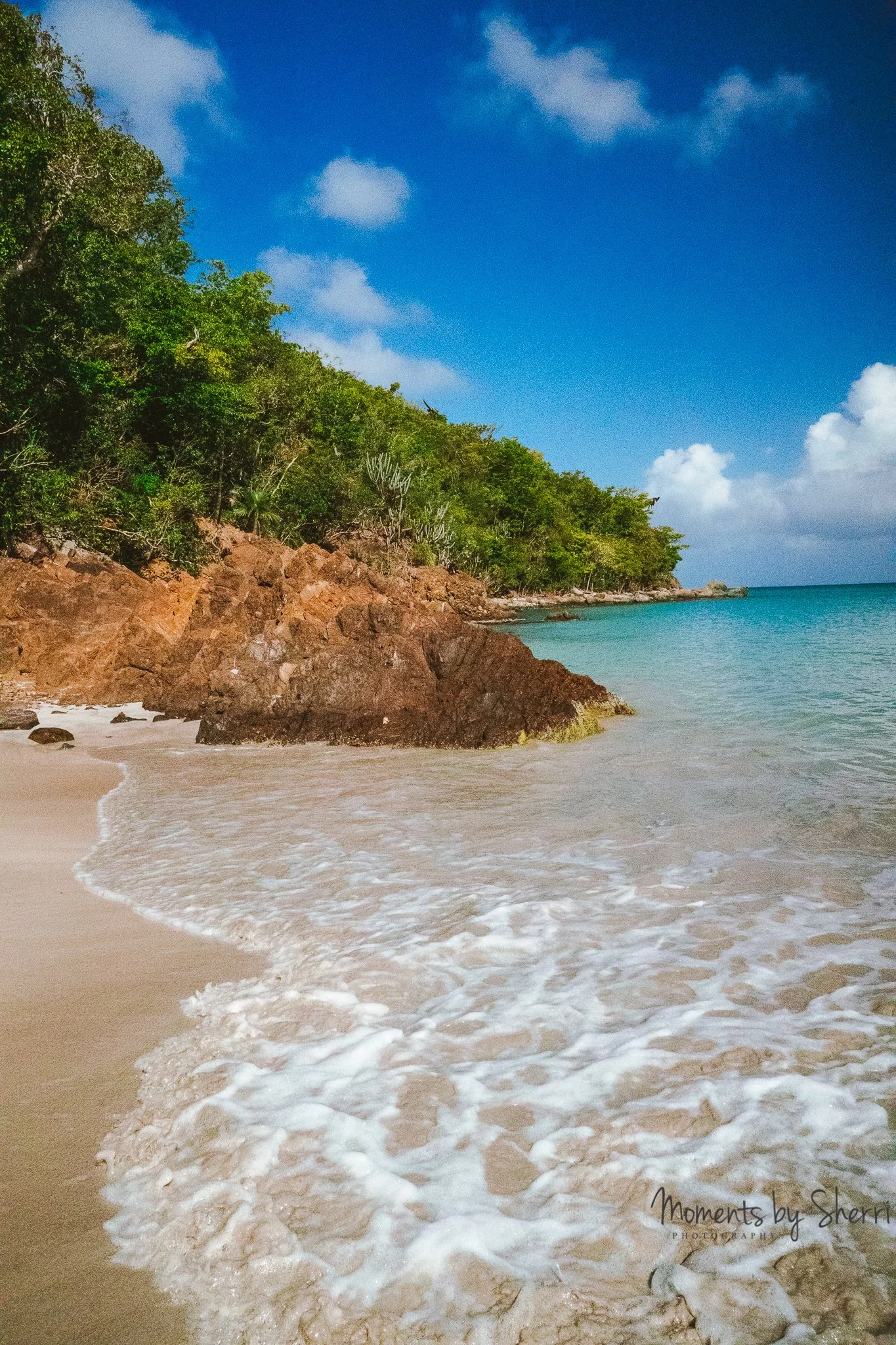 A beach with white sand, gentle waves, rocks, green trees on a cliff, blue sky with some clouds, and turquoise water.