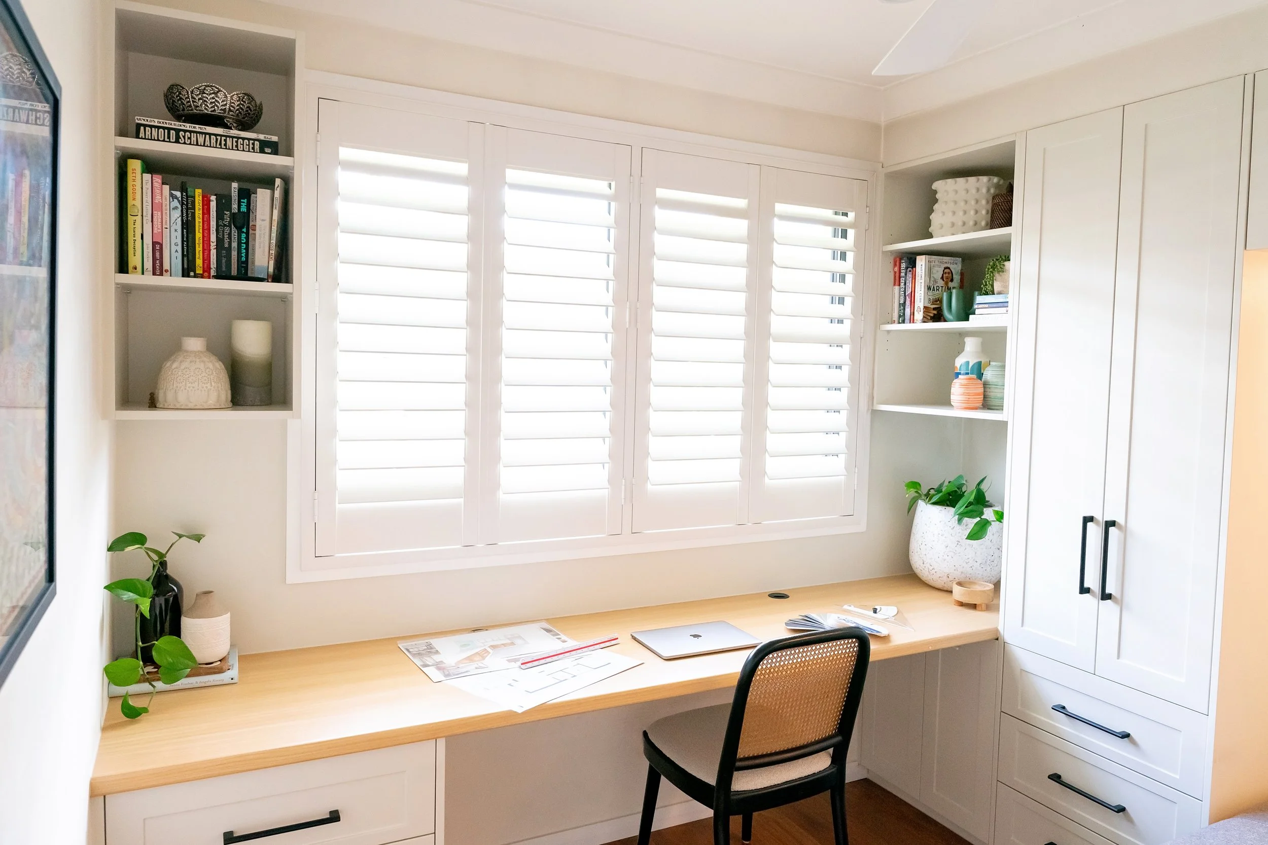 Home office with a wooden desk, black chair, white shelves, and a large window with white plantation shutters, decorated with books, potted plants, vases, and a few papers.