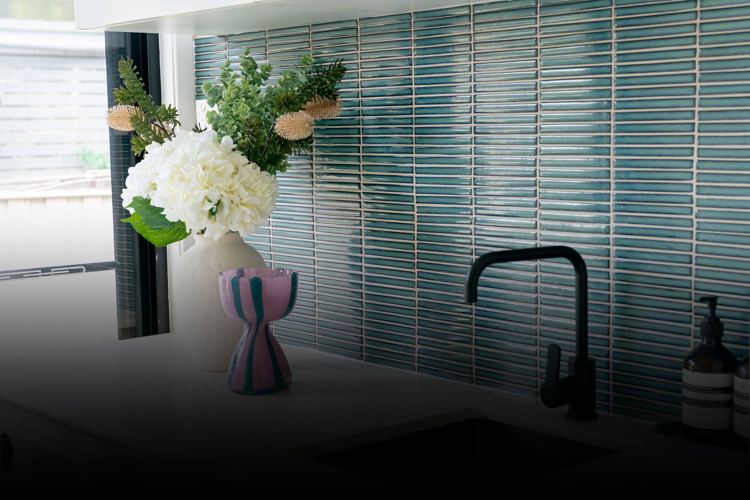 A kitchen countertop with a pink and purple vase holding a bouquet of white hydrangeas, green foliage, and artificial flowers, set against a backsplash of small horizontal blue tiles. A black faucet and soap dispenser are also visible.