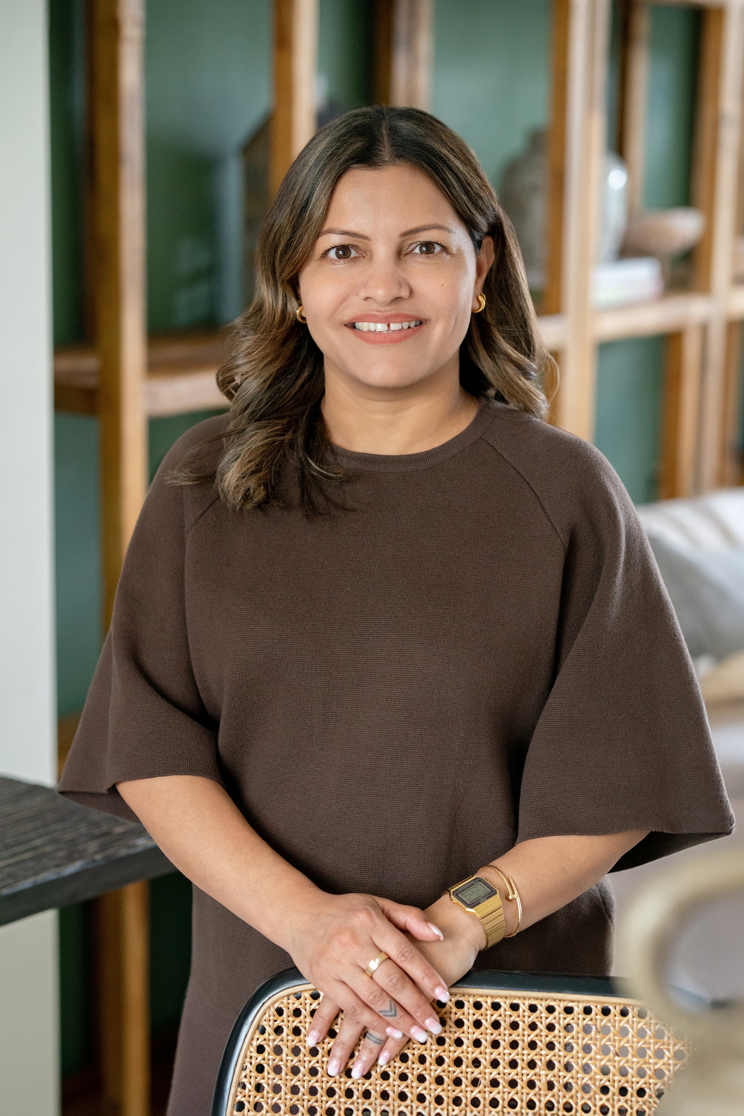 A woman with shoulder-length brown hair, wearing a brown top, gold earrings, a gold watch, and rings, standing indoors with wooden shelving and decorative objects in the background.