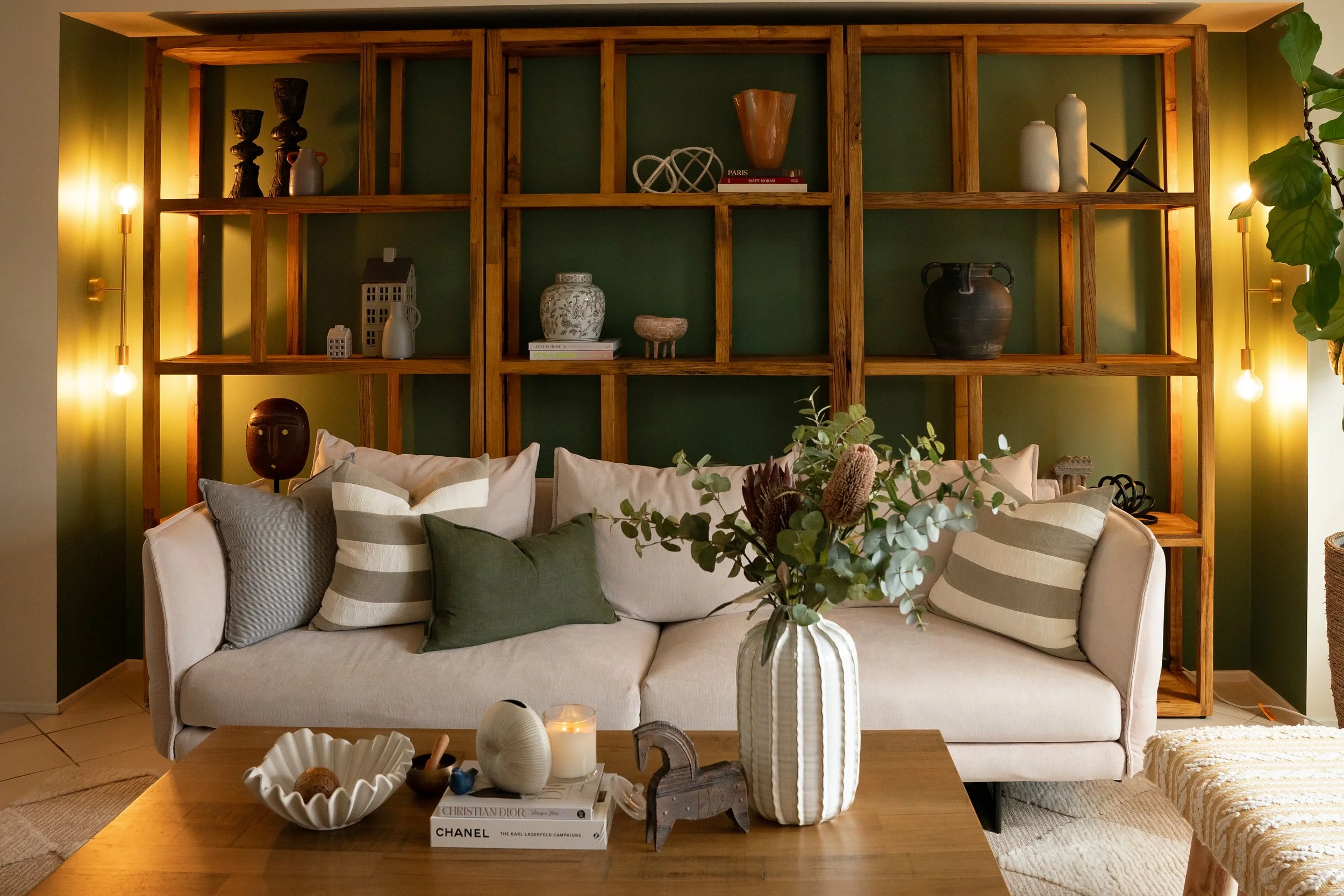 Living room with a beige sofa decorated with striped and plain pillows, a wooden coffee table with decorative items and books, a green accent wall with wooden wall shelves displaying vases, sculptures, and books, yellow wall-mounted lamps on either side of the shelves, and a large green leafy plant on the right side.