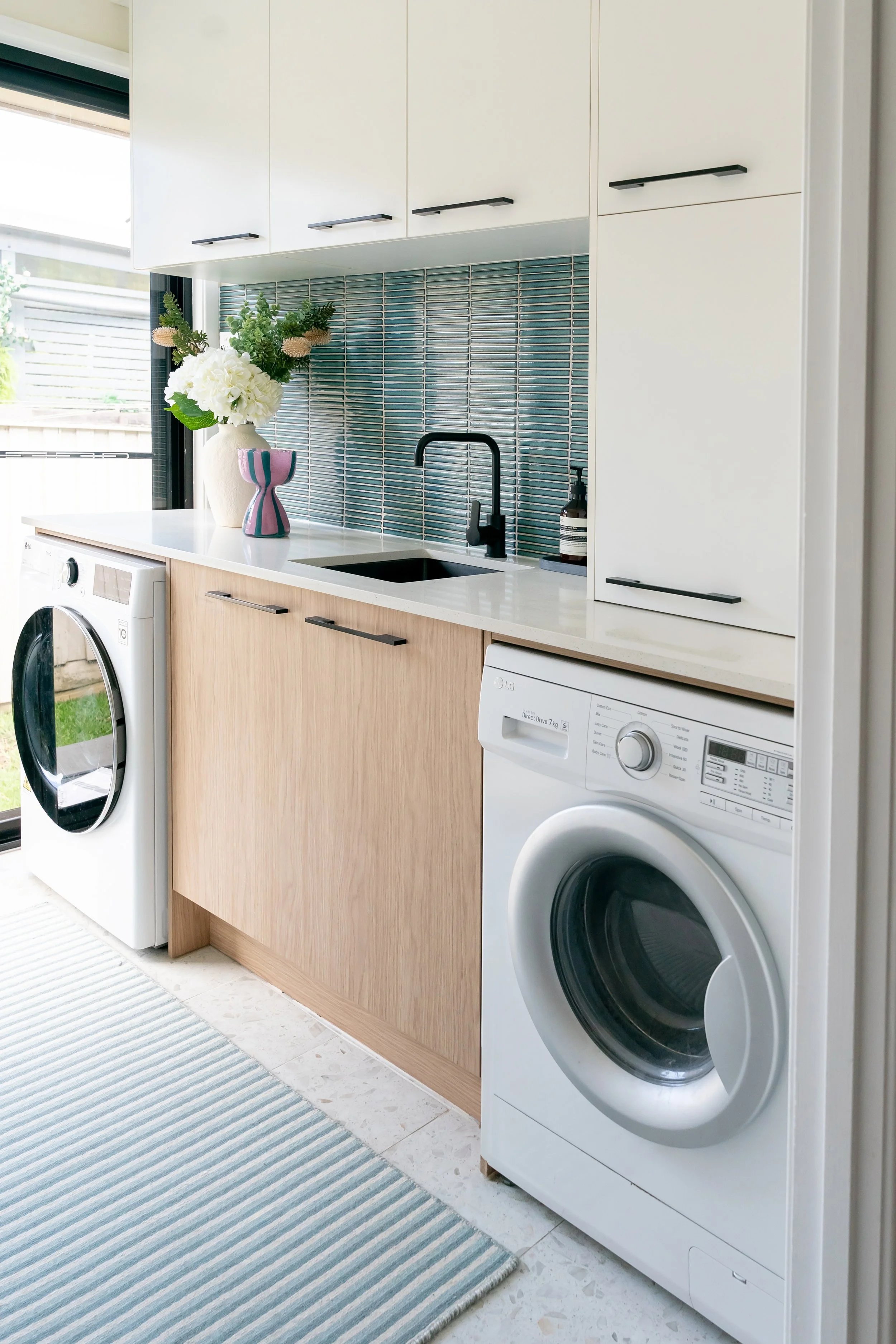 Laundry room with washing machine, sink, and white cabinets, decorated with a vase of white flowers and a pink and blue vase.