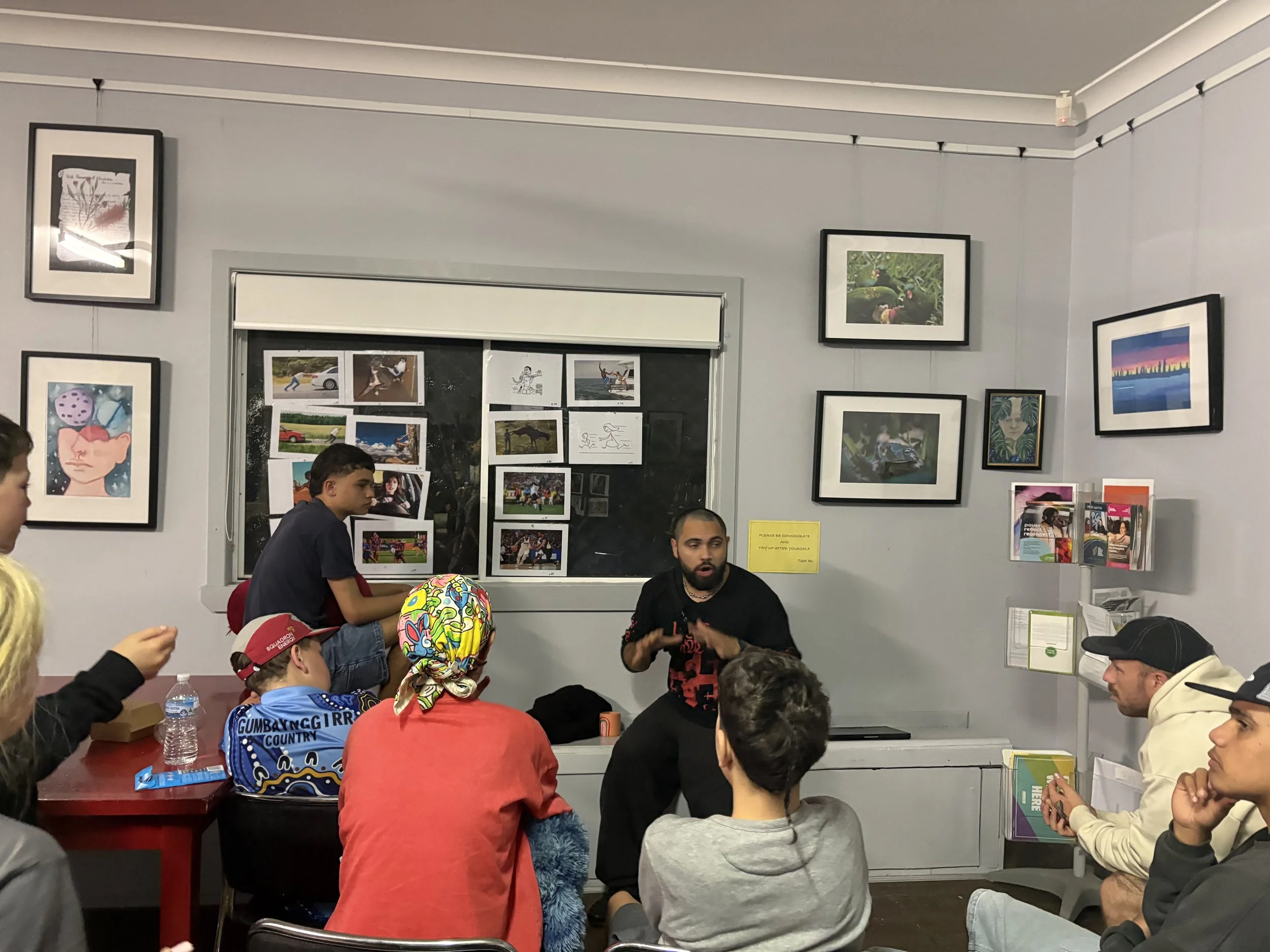 Birrugan Dunn-Velasco leading a group of people, including children and adults, gathered in a classroom or community room. Some individuals are standing while others are seated. Birrugan Dunn-Velasco leads the class.