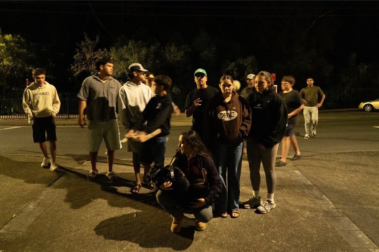 Photo taken by Birrugan Dunn-Velasco of group of young people standing and sitting outdoors at night, some wearing sweatshirts and hoodies, with trees and parked cars in the background. Founded by Birrugan Dunn-Velasco