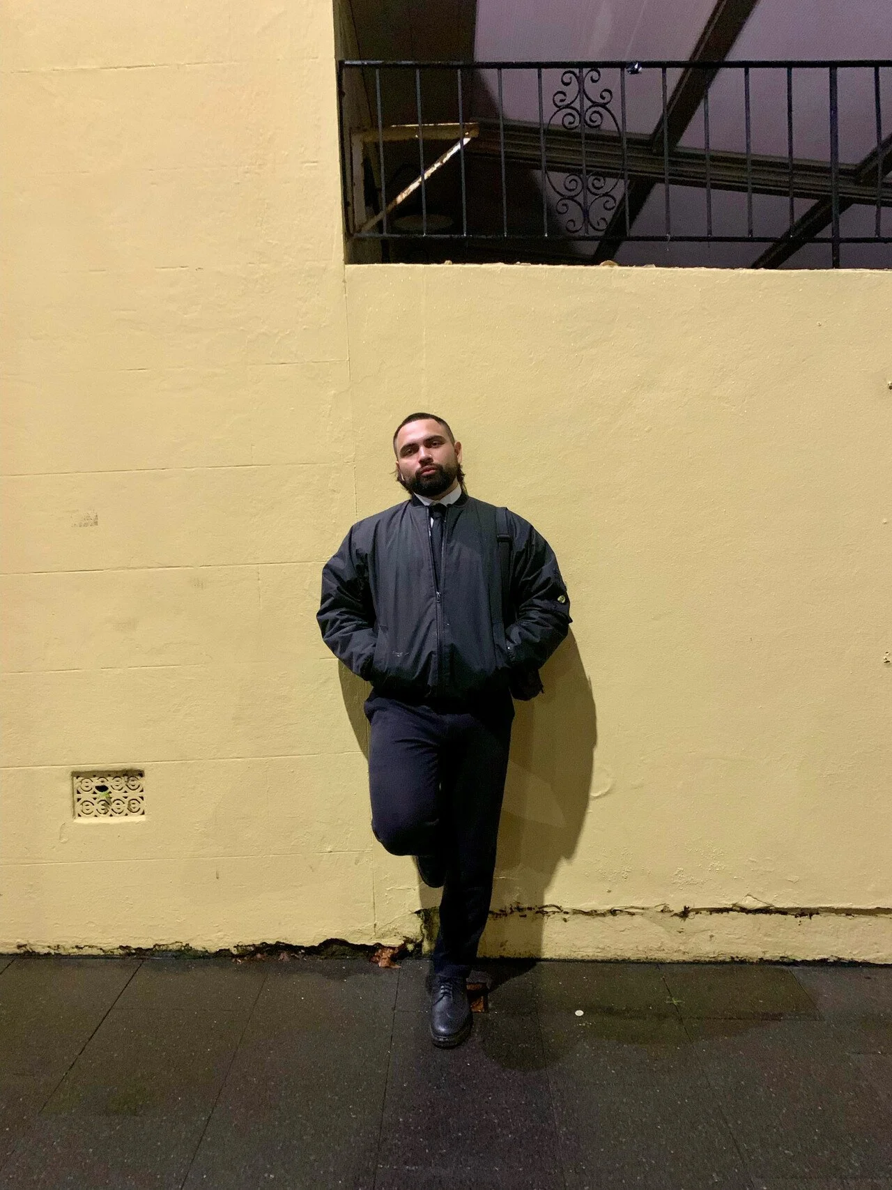 Birrugan Dunn-Velasco wearing a black jacket and black pants, standing against a yellow wall with one leg propped and his hands in his jacket pockets.