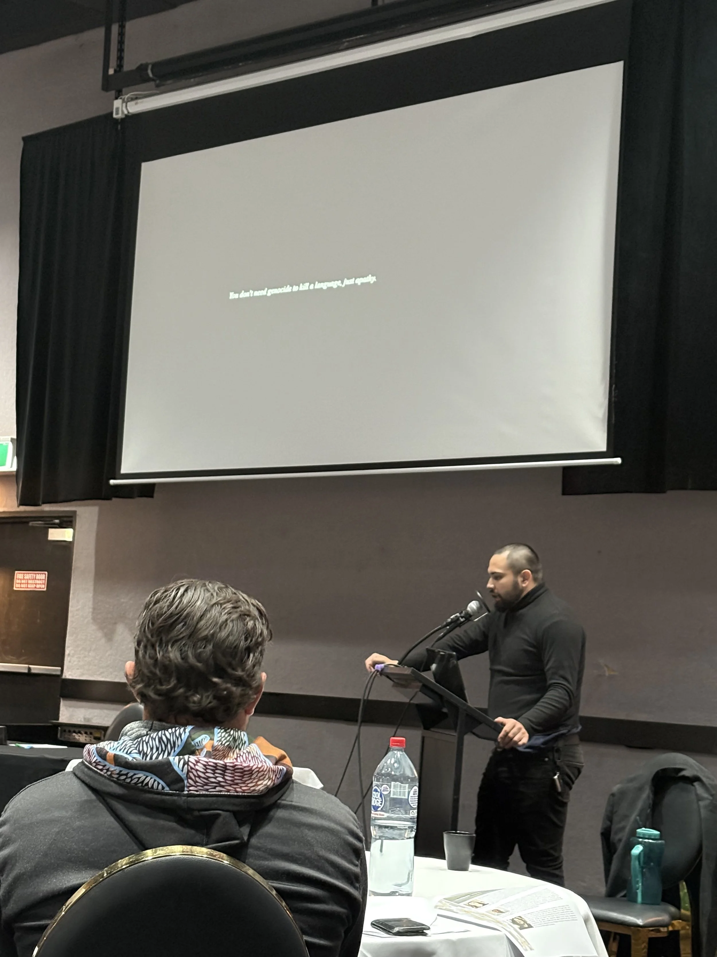 Birrugan Dunn-Velasco giving a presentation at a conference with a projected slide behind him. Several people are seated at round tables, listening. A large water bottle is on one table. The room has black curtains and dark walls.