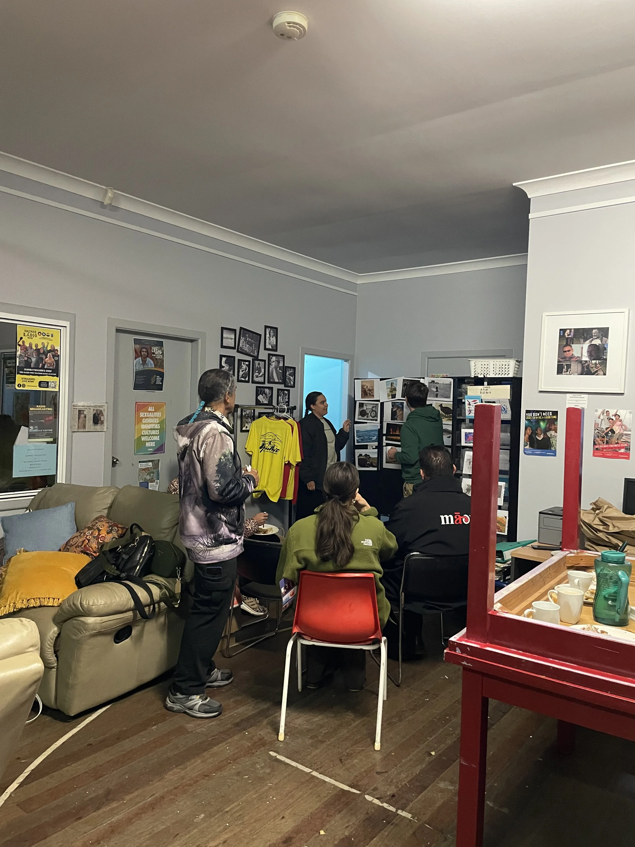 Group of people gathered in a room with framed photos on the walls, a white door, calendar, and display boards. Some seated, some standing, with food and mugs on a table in the foreground. Mililma May teaching. 