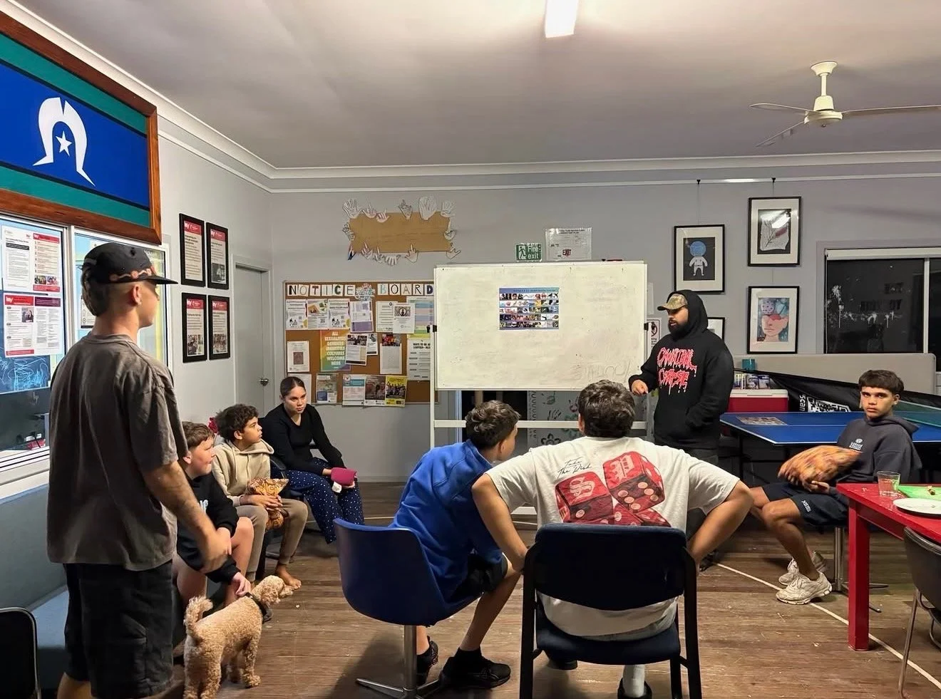 A group of children and a young man in a room with notice boards, framed pictures, a whiteboard, and a ping pong table. One child has a small dog sitting beside him. The children are seated on chairs and benches, facing a young man who appears to be speaking or giving instructions. The room has a ceiling fan and artistic framed photos on the wall.