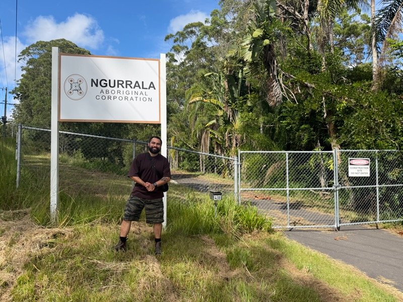 Birrugan Dunn-Velasco stands smiling in front of a fence with a sign for Ngurrala Aboriginal Corporation at the entrance of a wooded area.