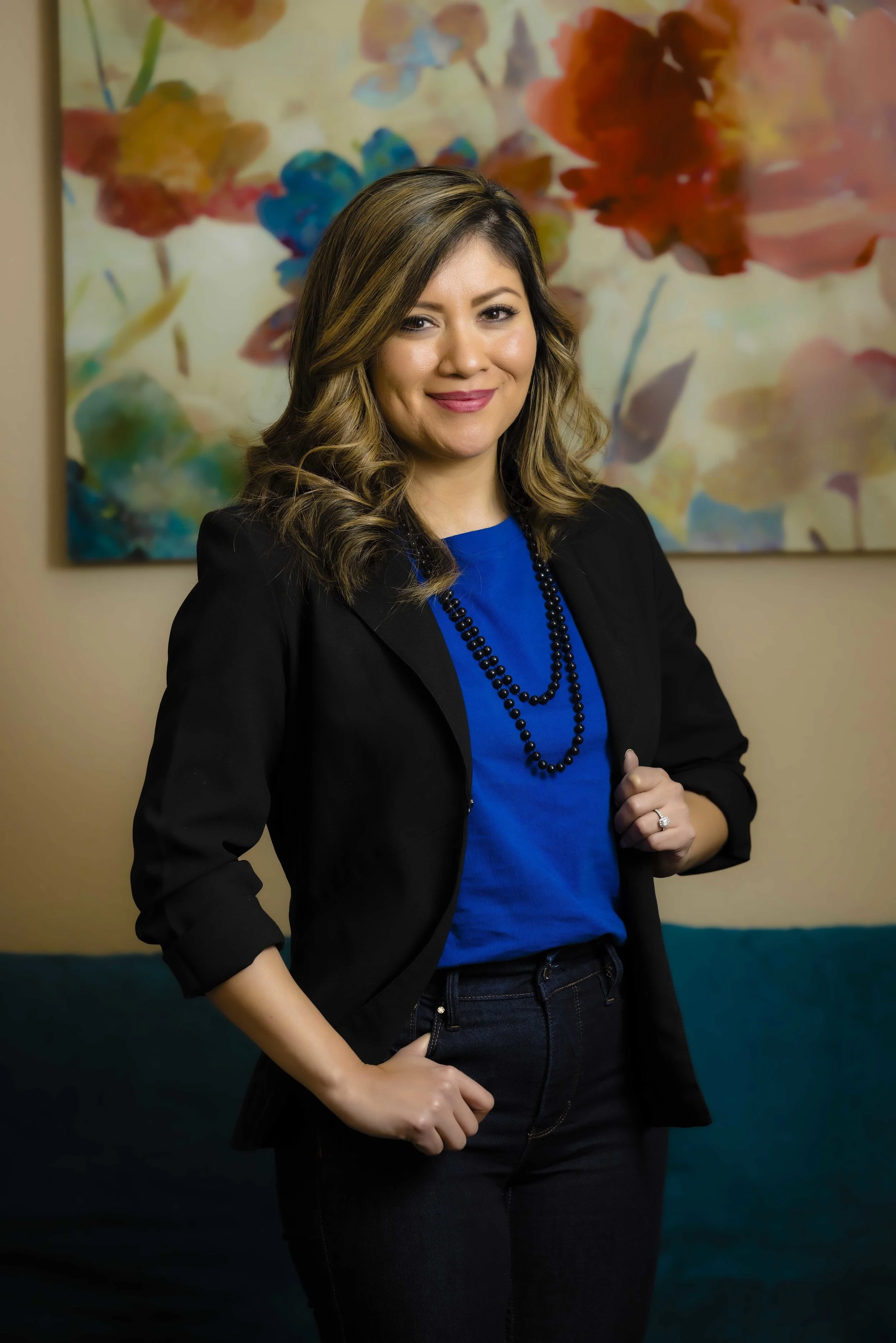 A woman with wavy, shoulder-length light brown hair, smiling, wearing a blue top, black blazer, black beaded necklace, and black jeans, standing in front of an abstract floral painting background.