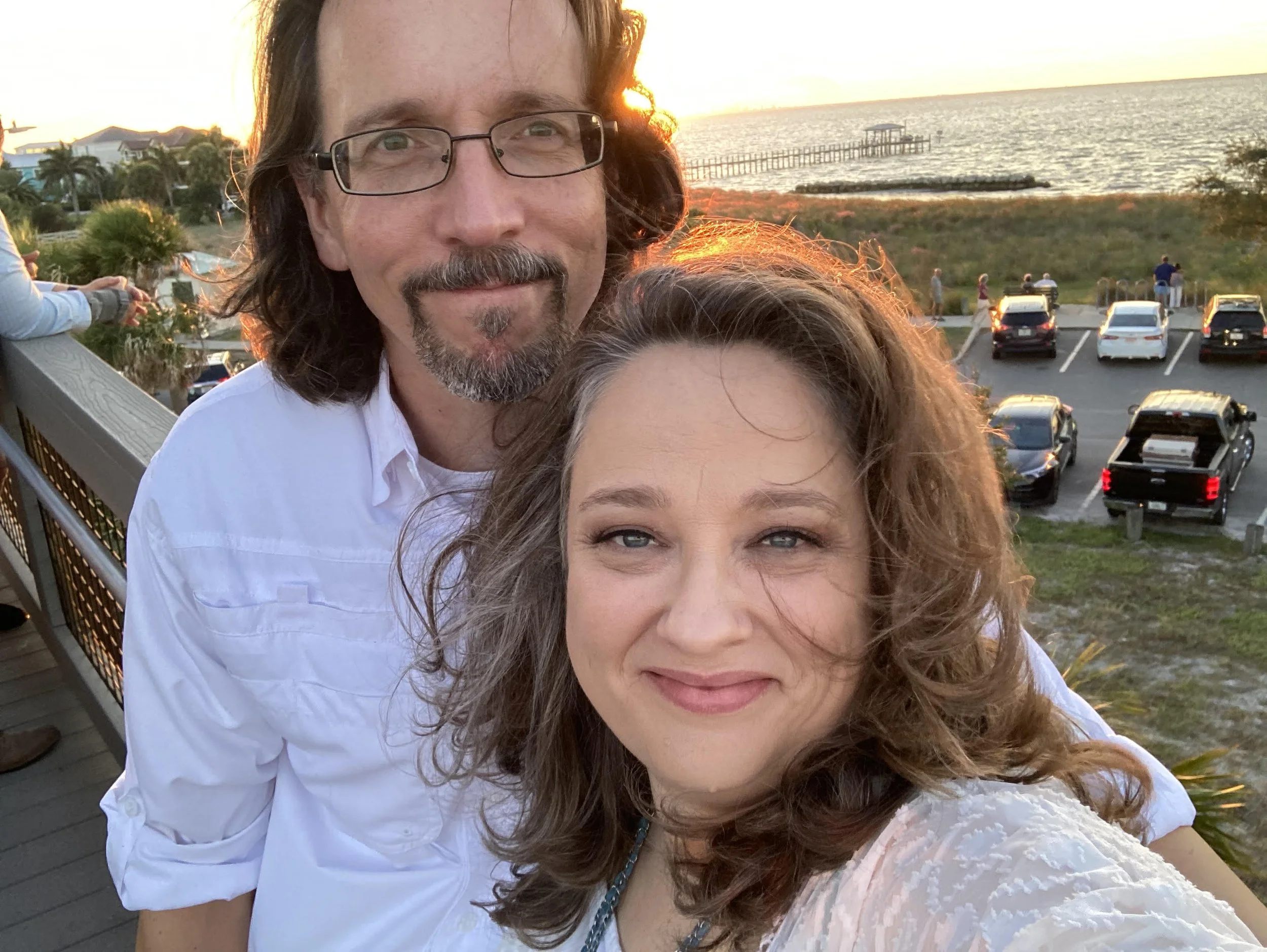 A man and woman taking a selfie on a balcony overlooking a parking lot, a beach, and a pier at sunset.