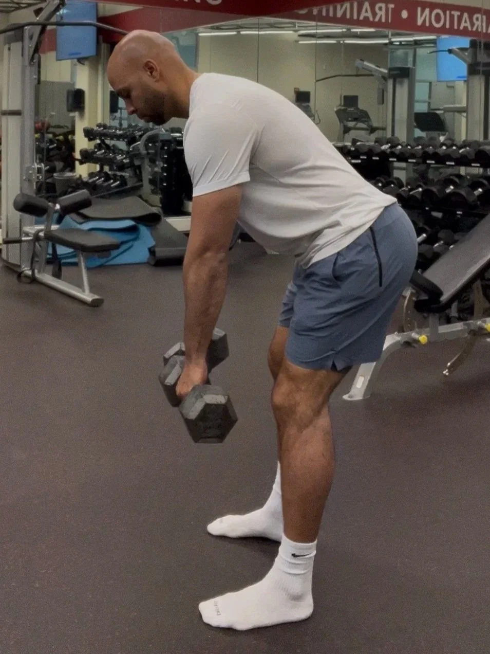 A man lifting dumbbells in a gym, bending at his hips with a flat back, wearing a white t-shirt, blue shorts, and white Nike socks.