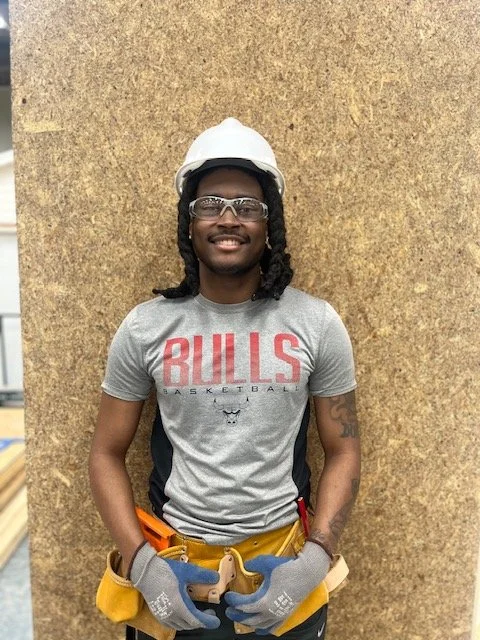 A young man wearing safety goggles, a white hard hat, work gloves, and a Bulls basketball T-shirt, standing against a cork board wall.