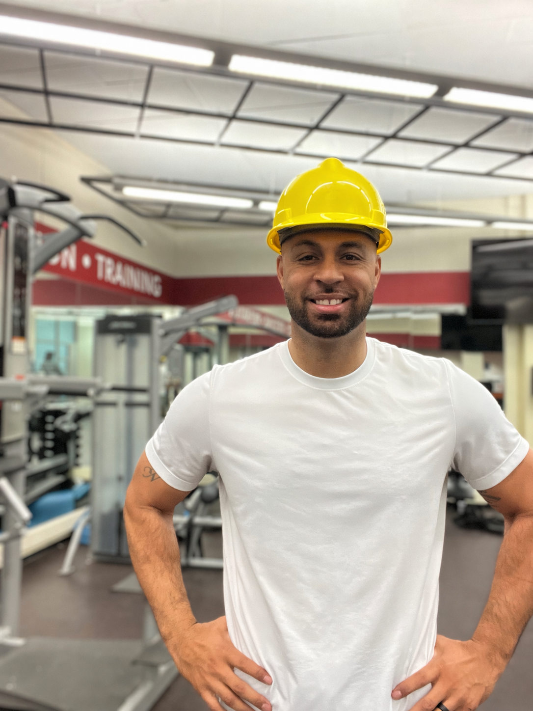A man smiling and wearing a yellow safety helmet inside a gym with weightlifting equipment and a sign that reads 'TRAINING' in the background.