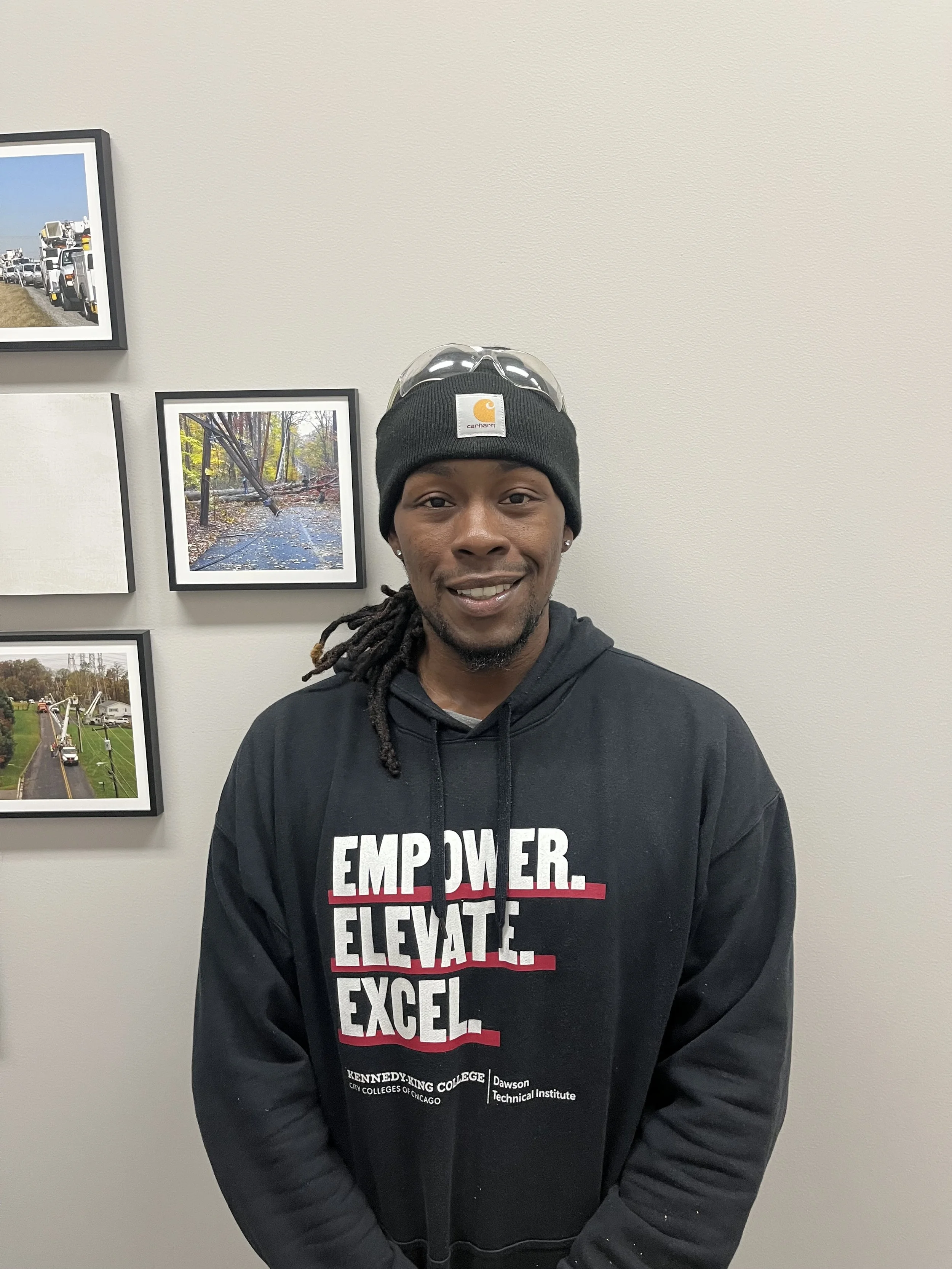 A smiling man with dreadlocks wearing a black beanie, clear safety glasses on top of his beanie, and a black hoodie with white and red text that says 'Empower. Elevate. Excel.' standing in front of a white wall with framed photos of trucks and a forest scene.