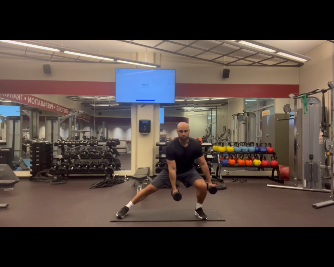 Man performing a side lunge exercise with dumbbells in a gym, surrounded by weights and fitness equipment.