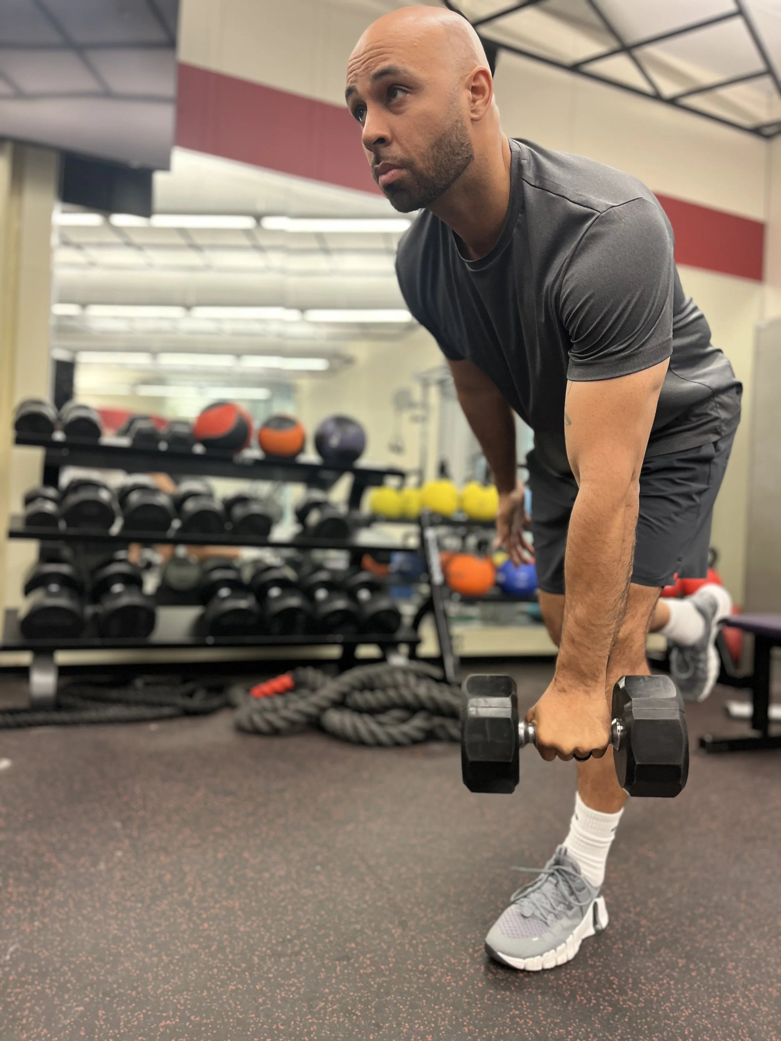 A man with a bald head, beard, and gray athletic clothing performing a single-leg deadlift exercise with a dumbbell in a gym, with storage racks of weights and medicine balls in the background.