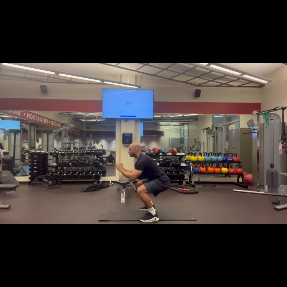 Man performing squat exercise on a mat in a gym with various exercise equipment and colorful kettlebells.