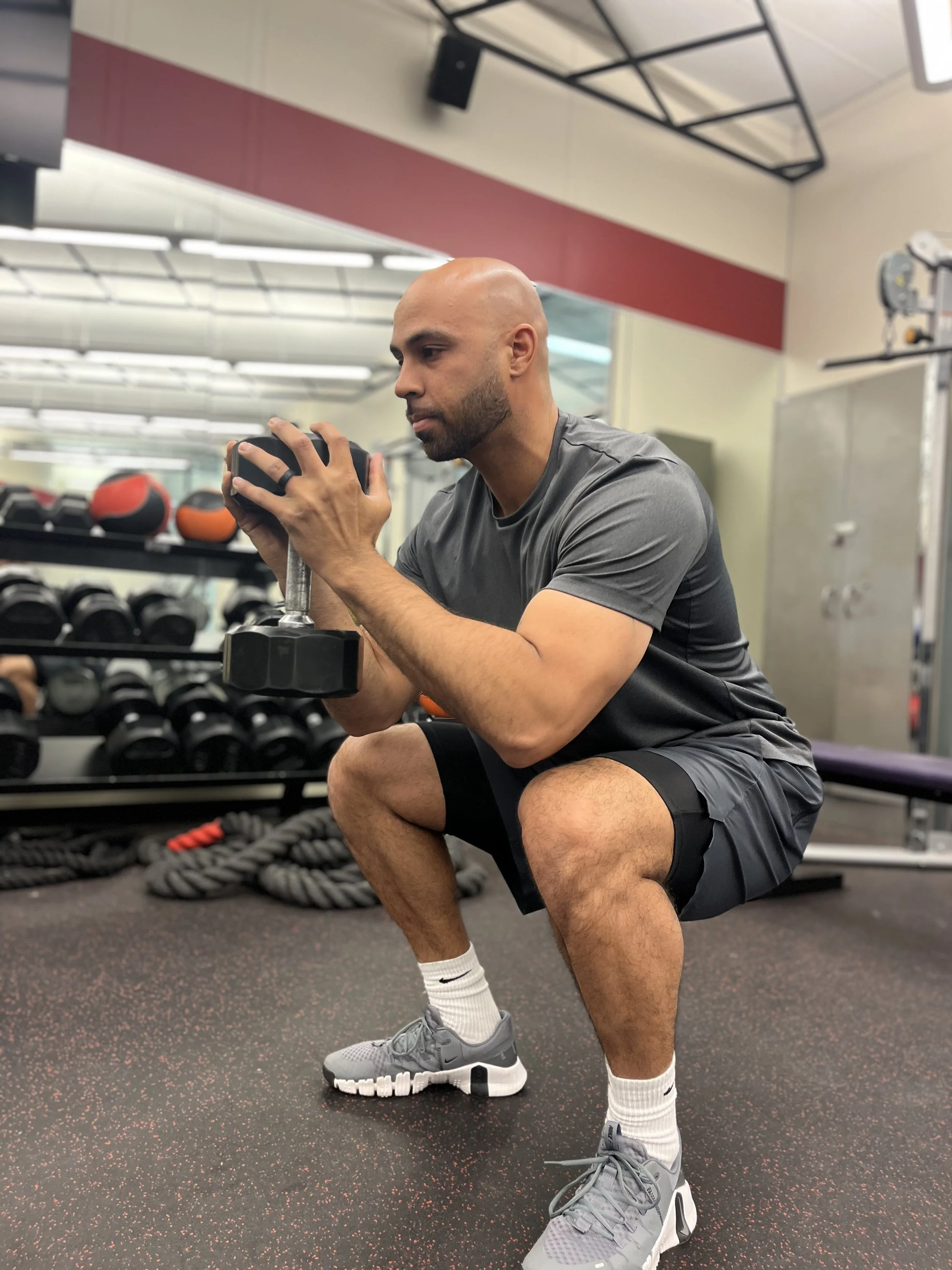 A man performing a squat exercise with a dumbbell in a gym. He is wearing gray workout clothes and sneakers, with equipment and weights in the background.