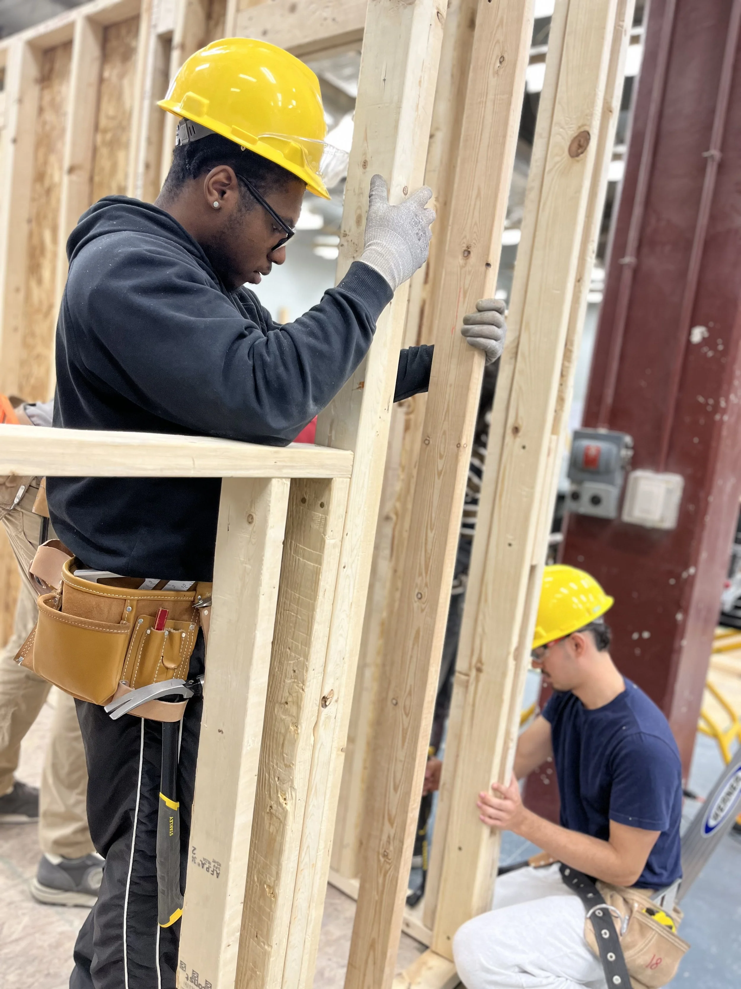 Two construction workers wearing yellow safety helmets and working on wooden frames.