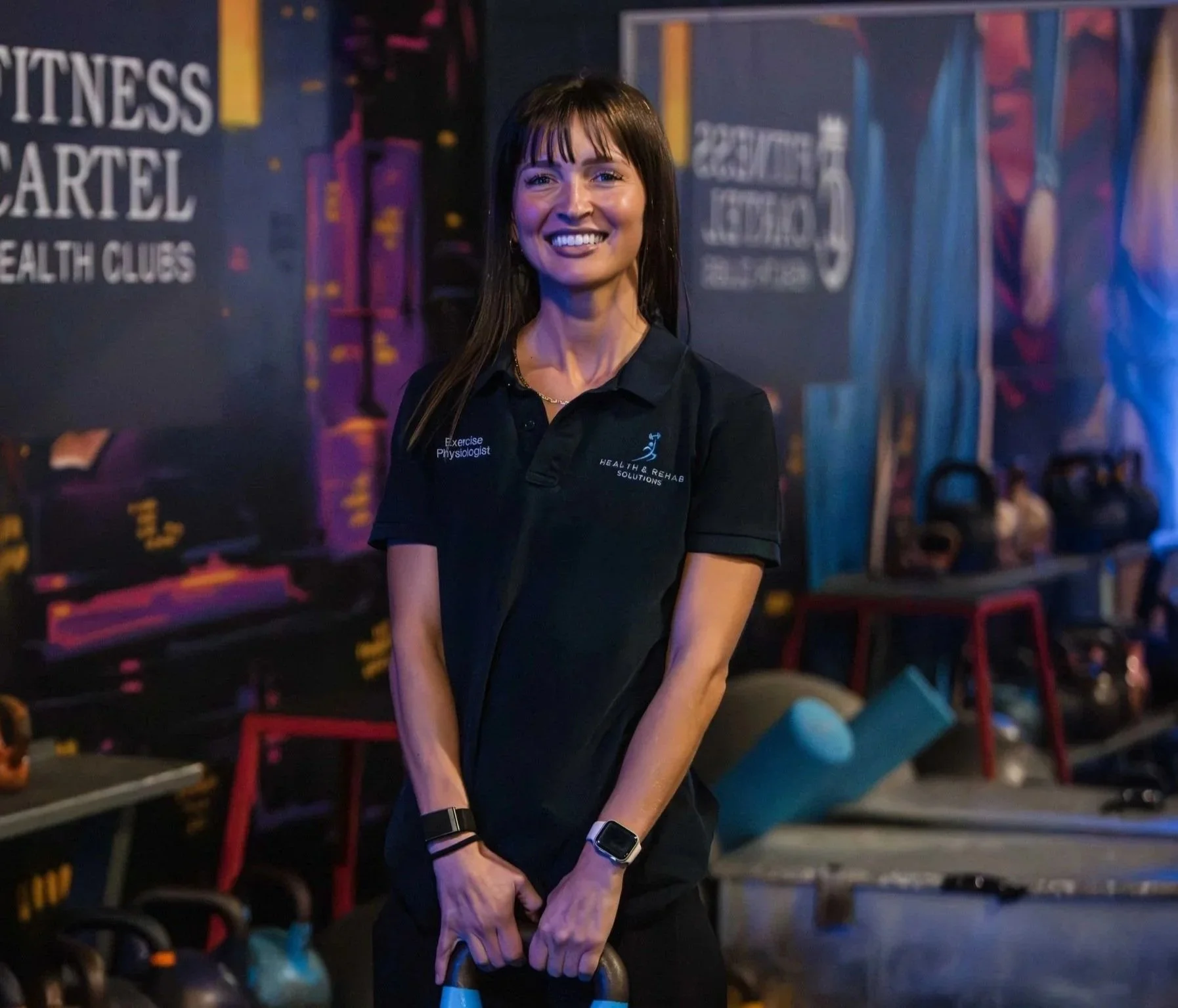 Woman smiling at a gym, wearing a black polo shirt with the words 'Exercise Physiologist' and 'Health & Rehab Solutions'. She is holding a kettlebell and is surrounded by gym equipment and motivational posters.
