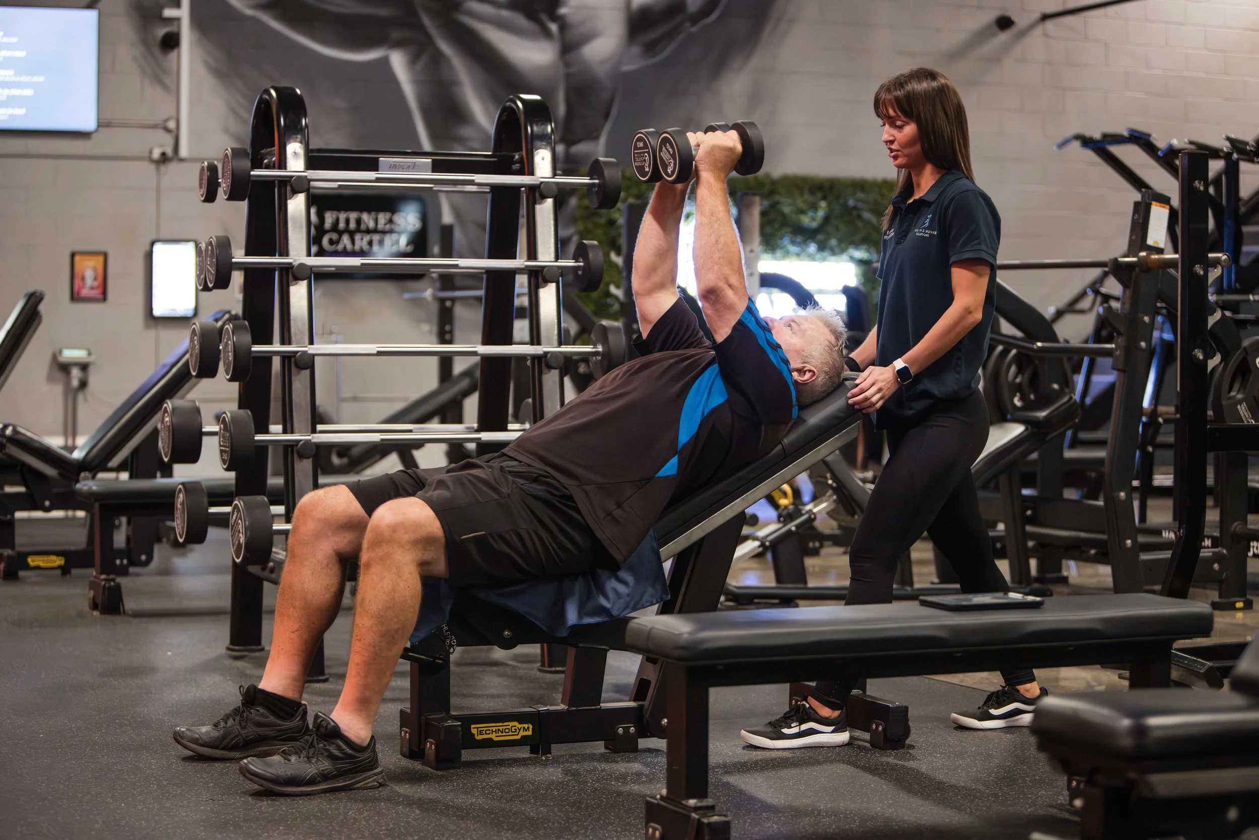 An elderly man lying on a workout bench doing a dumbbell chest press exercise with a trainer standing nearby in a gym. The gym has various workout equipment and a sign that reads 'Fitness Cakie' in the background.
