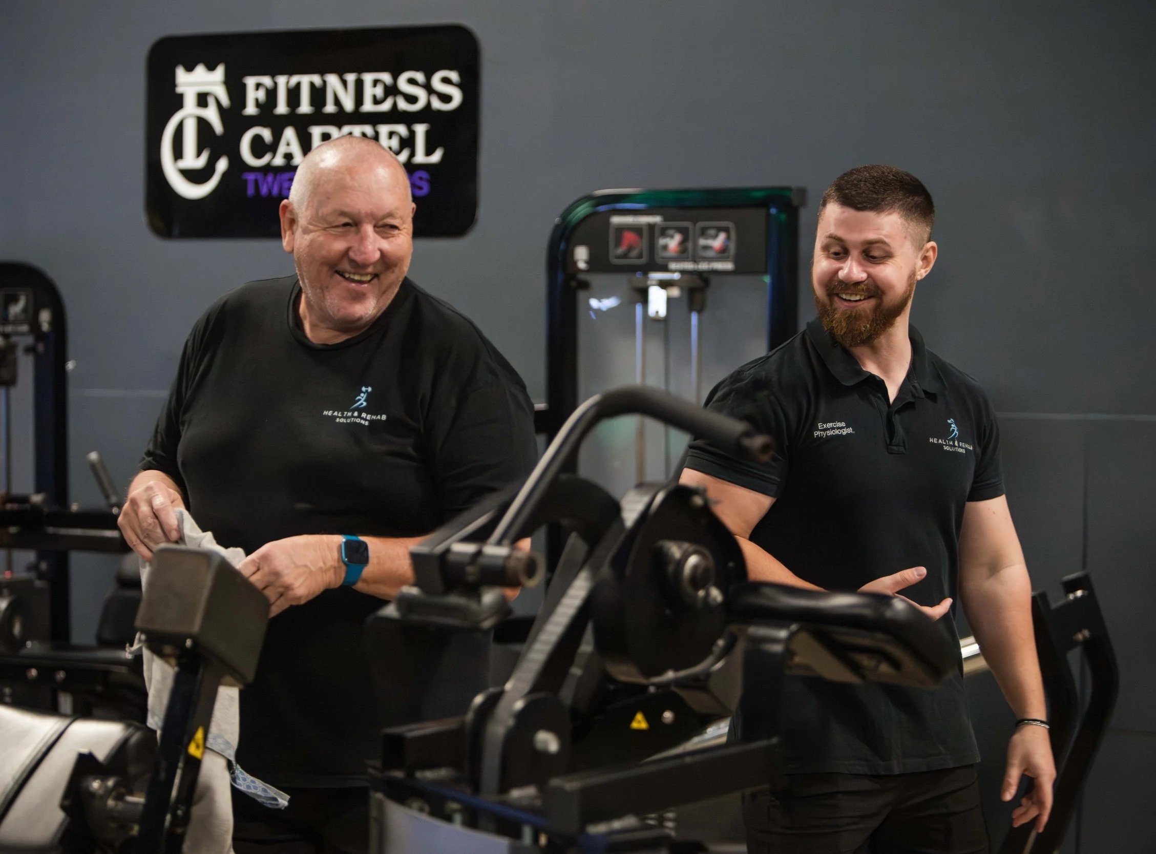 Two men smiling and engaging in conversation at a gym, with exercise equipment visible in the foreground. One man is older, with a bald head, wearing a black T-shirt with a logo, and the other is younger, with a beard, also wearing a black shirt. A sign in the background reads 'Fitness Barrel'.