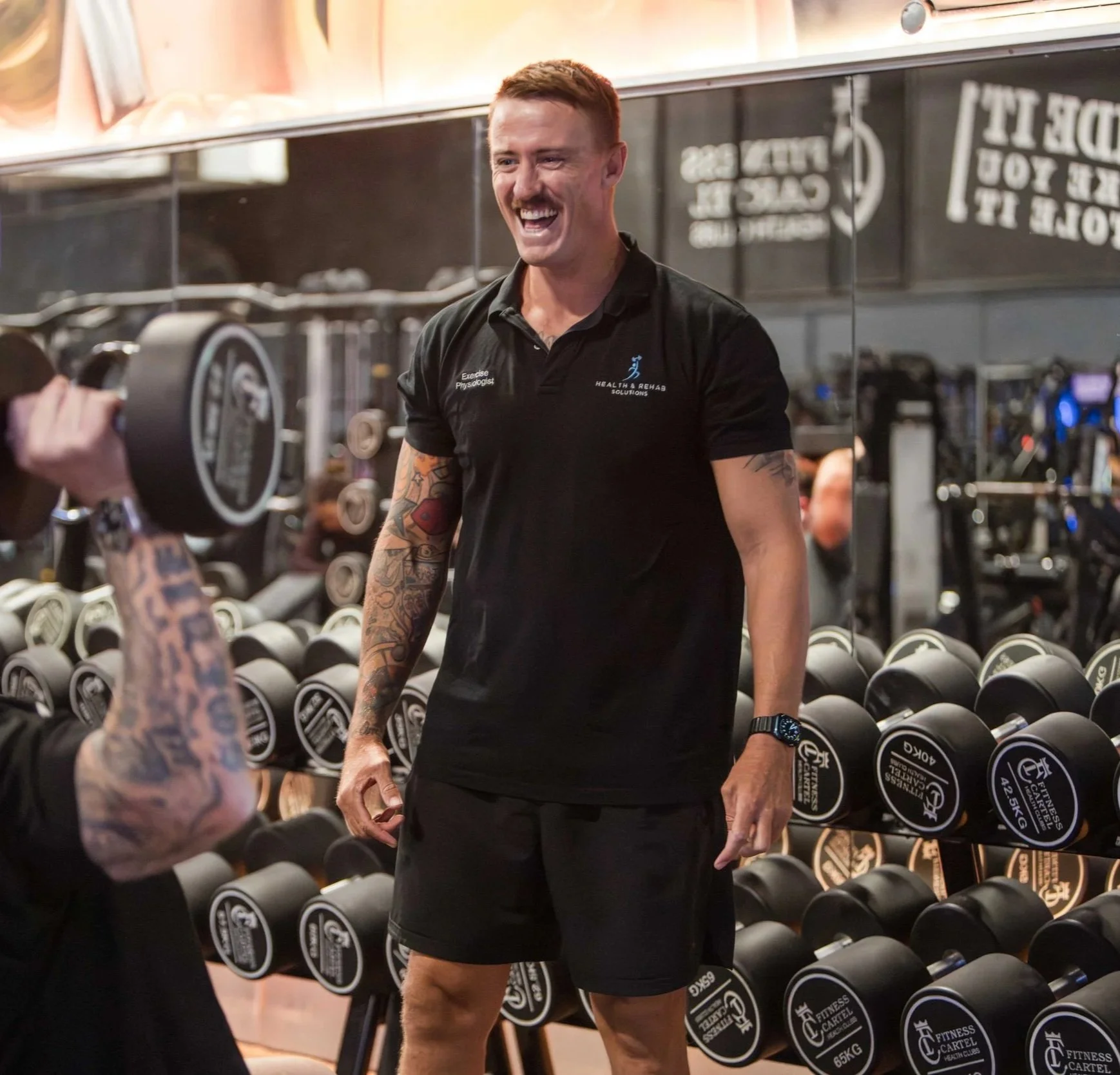 A man with red hair and tattoos on his arm is smiling in a gym, standing in front of a rack of dumbbells. He is wearing a black polo shirt with a logo and text that says 'Health & Rehab Solutions' and 'Exercise Physiologist'. He appears to be interacting with another person who is holding a camera.
