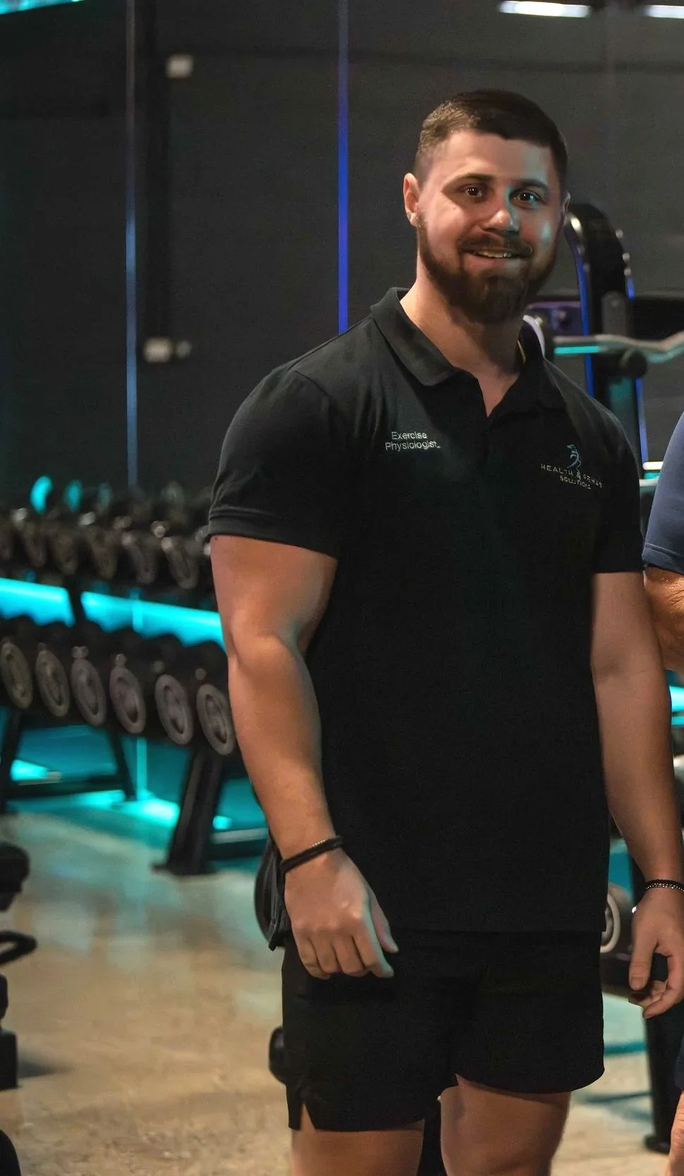 Man with short hair and beard in a gym, wearing a black polo shirt and black shorts, standing near exercise equipment.