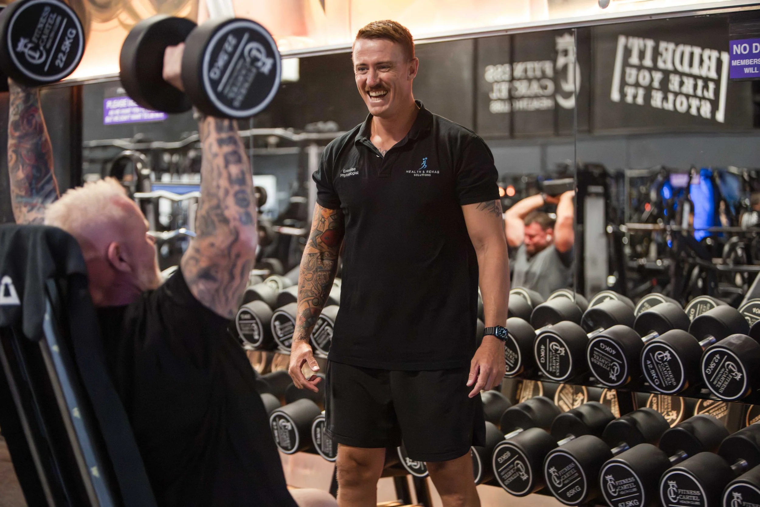 A man with tattoos wearing a black fitness shirt and shorts smiling and talking to an elderly person with tattoos in a gym, with rows of dumbbells and a mirror behind them.