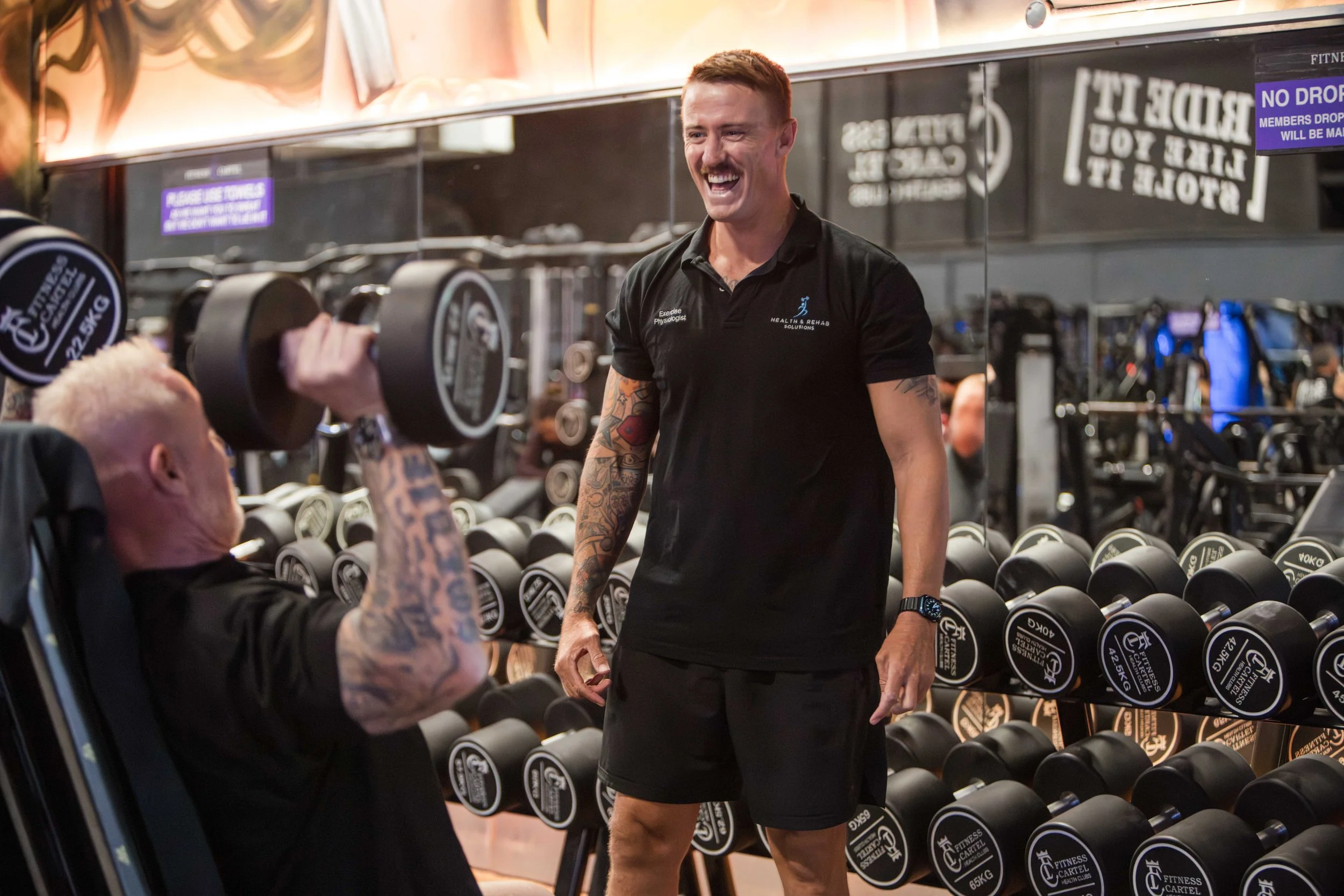 A man smiling and talking to a person lifting dumbbells in a gym. The man has tattoos on his arms, short hair, and is wearing a black polo shirt with “Exercise Physiologist” and “Health & Rehab Solutions” written on it.