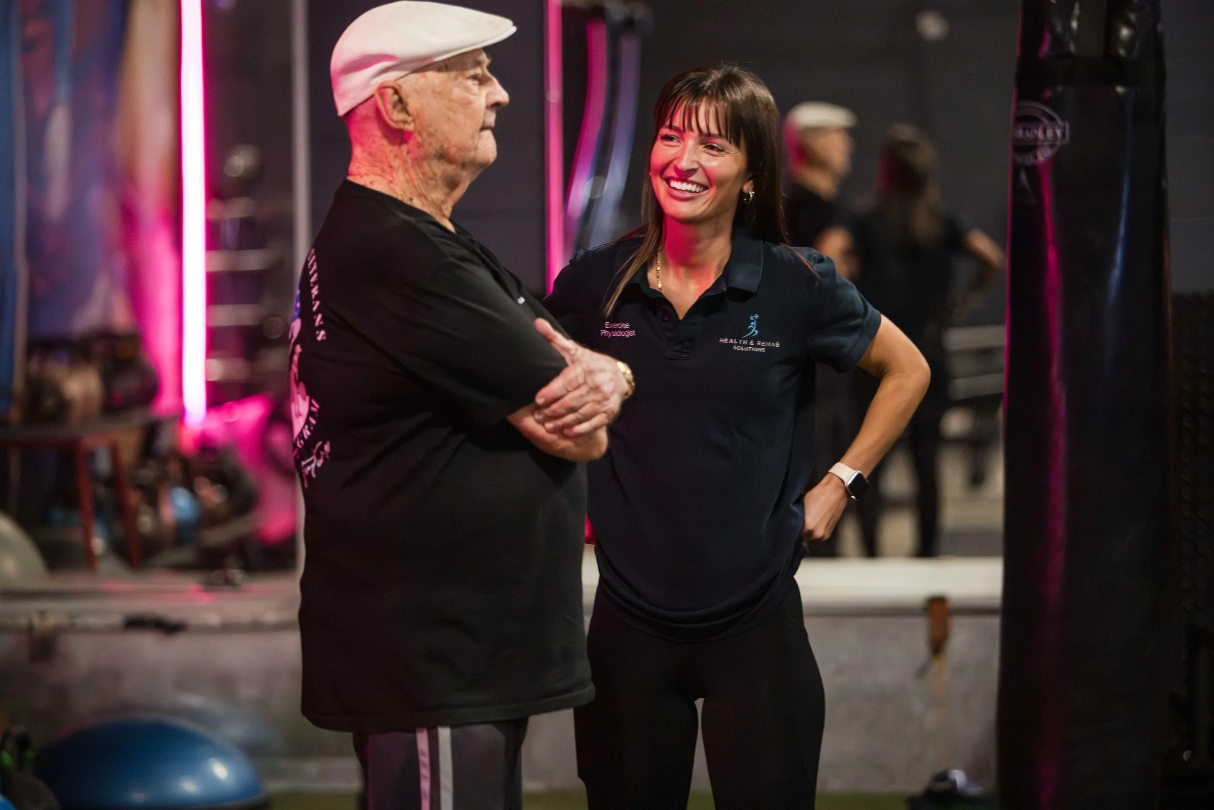 A fitness instructor and an older man talking in a gym. The instructor is smiling and wearing a navy polo shirt with 'Exercise Physiologist' and 'HEALTH & REHAB SOLUTIONS' printed on it. The man is wearing a white cap, black shirt, and appears to be engaged in conversation with the instructor.