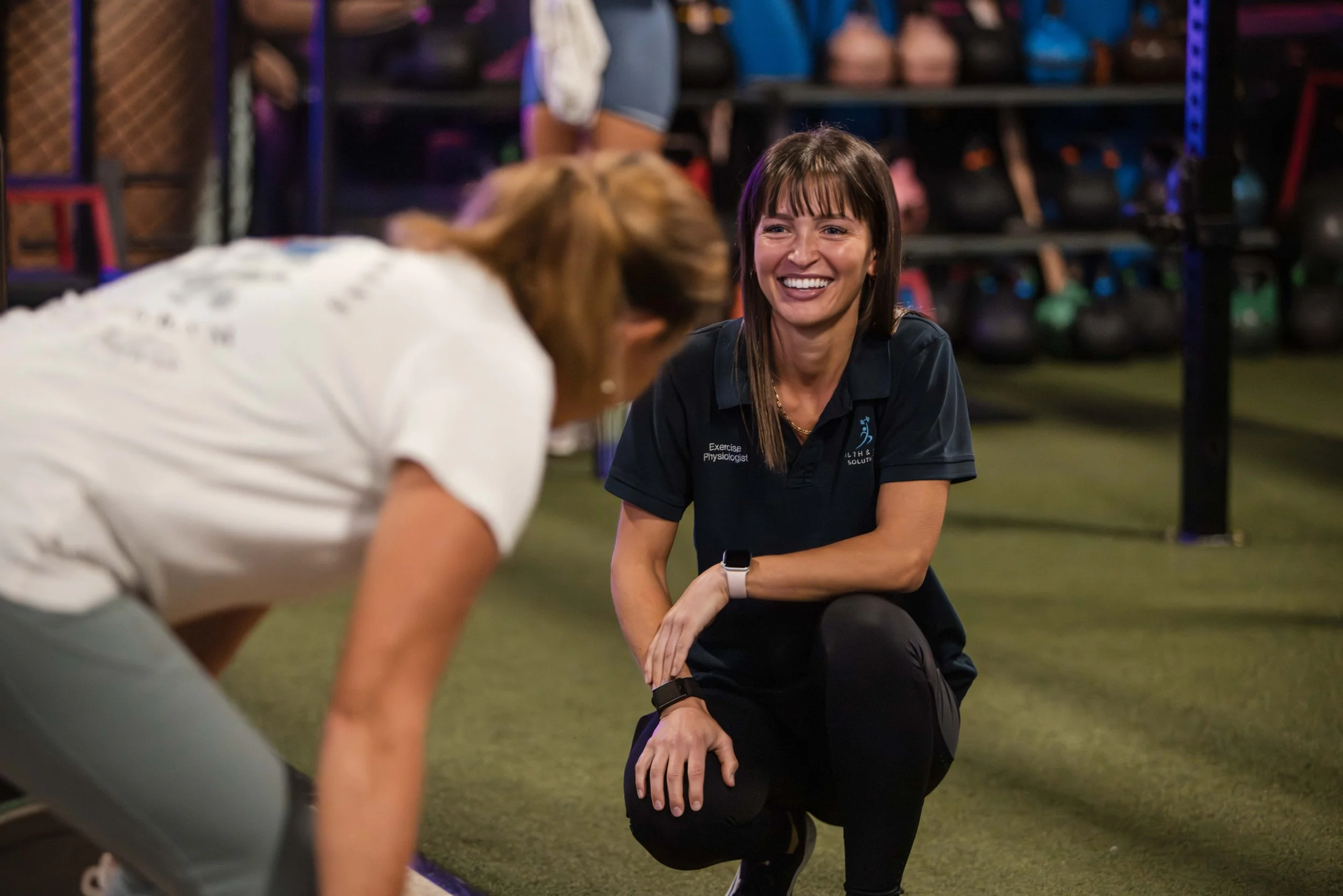 A female exercise physiologist with brown hair, wearing a black shirt with name tag, smiling and coaching a woman during a workout in a gym with weights and kettlebells in the background.