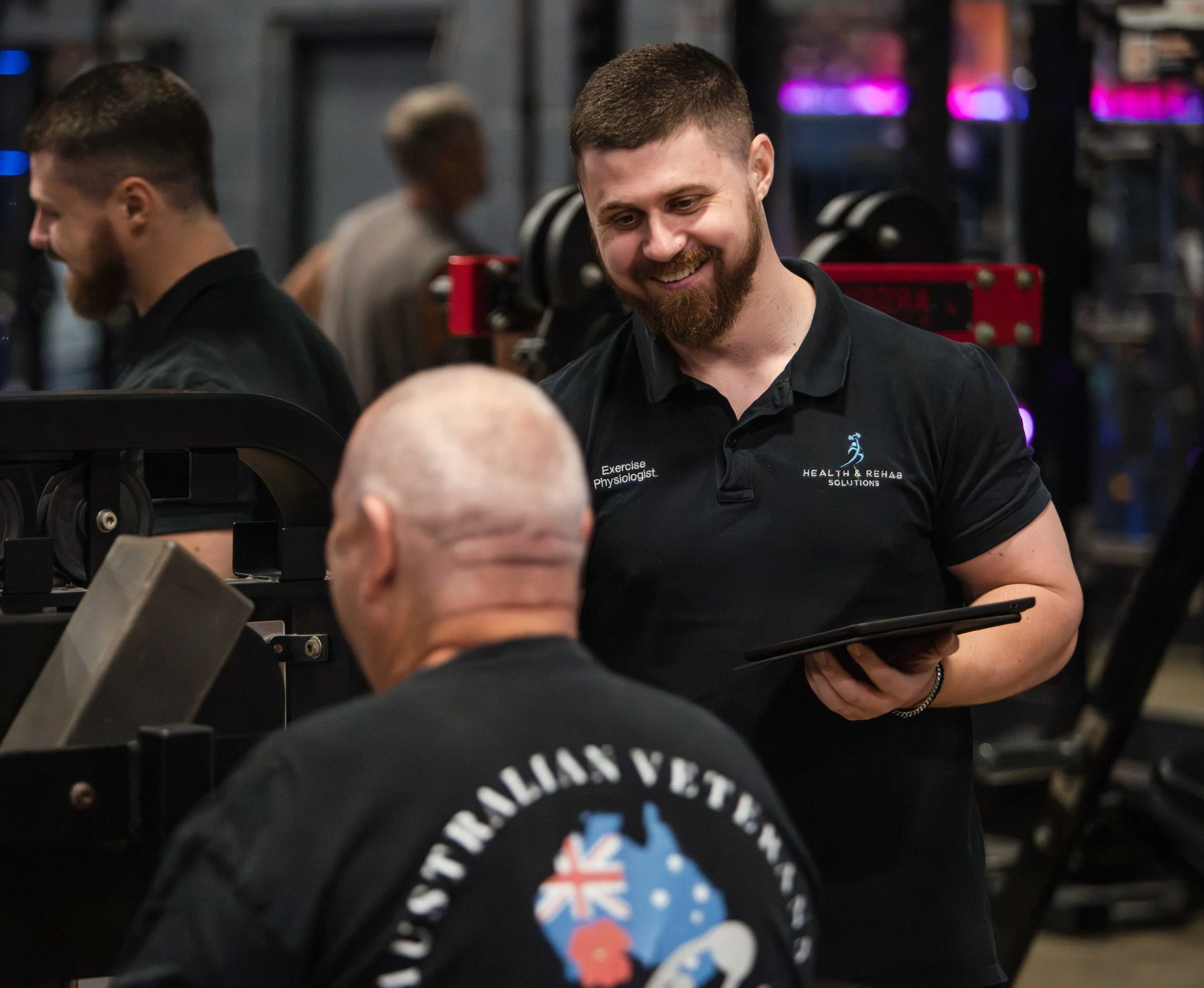 Two men with beards and short hair talking and smiling; they are in a gym or fitness center with equipment in the background; one is wearing a black athletic shirt with a logo and text, the other has dark hair and a beard.
