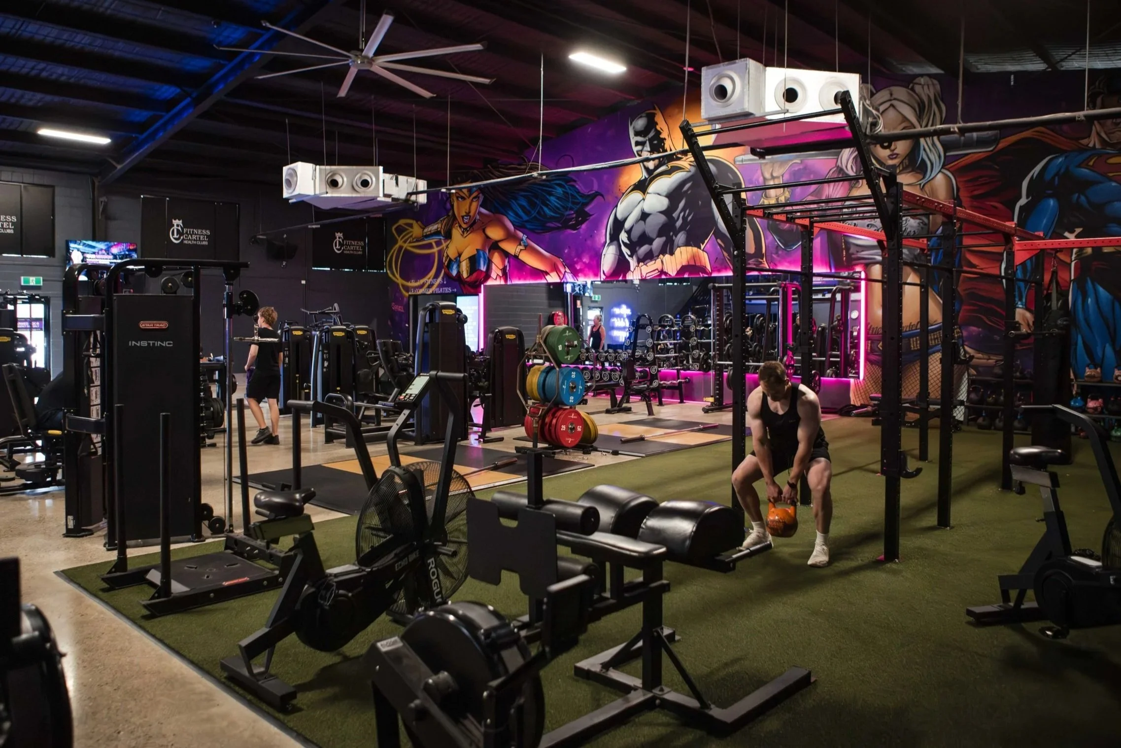 Inside a gym with workout equipment, colorful wall murals featuring comic-style characters, and people working out.