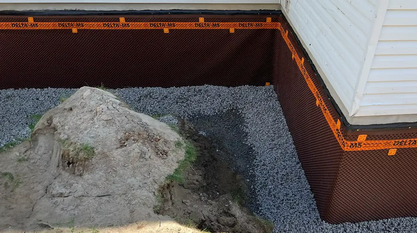Travaux de fondation avec panneau d'étanchéité autour de la base d'une maison, avec des gravillons et une pierre large au premier plan.