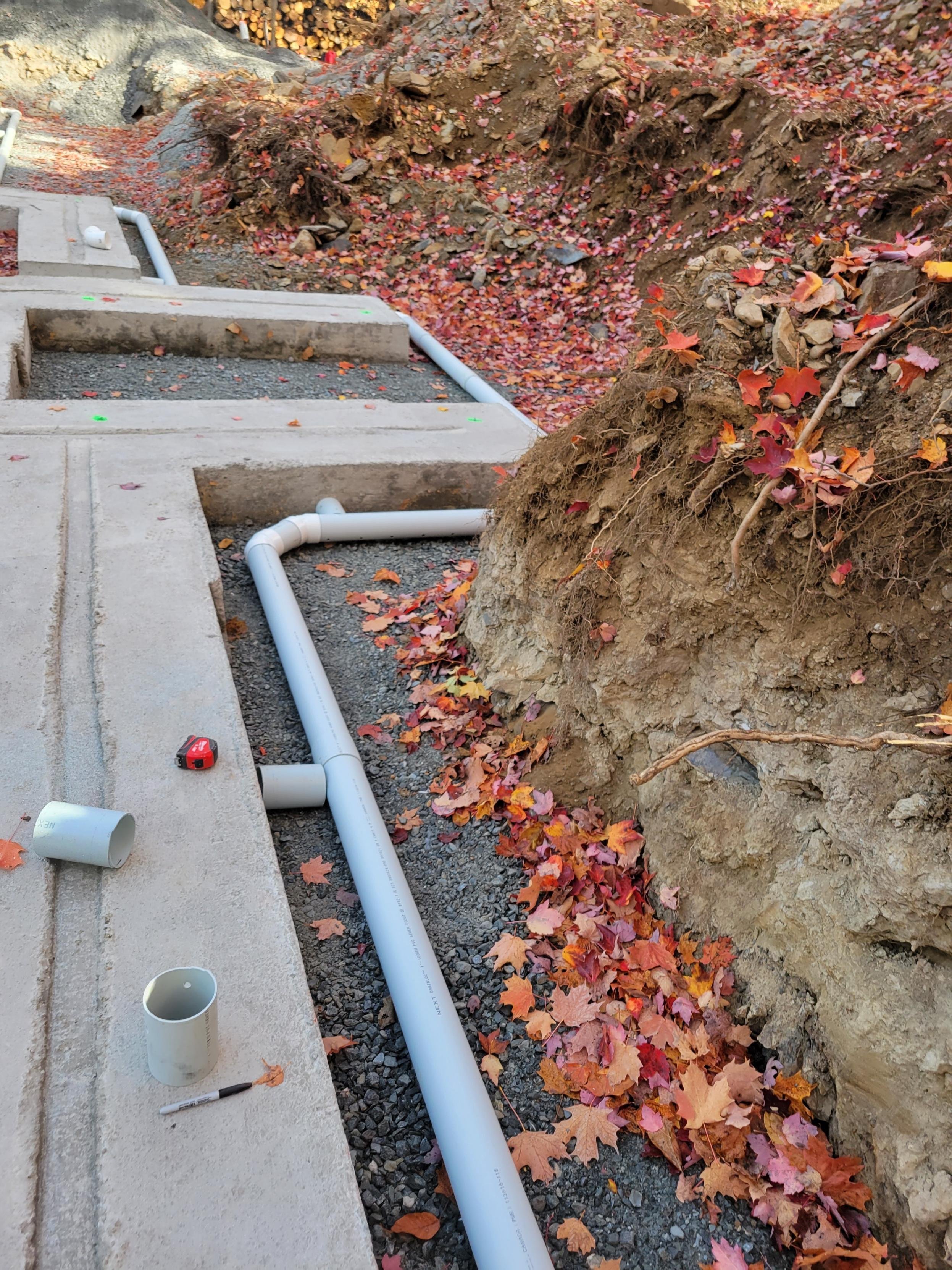 Travaux de plomberie en cours avec des tuyaux en PVC dans un sol en terre entouré de feuilles mortes automnales.