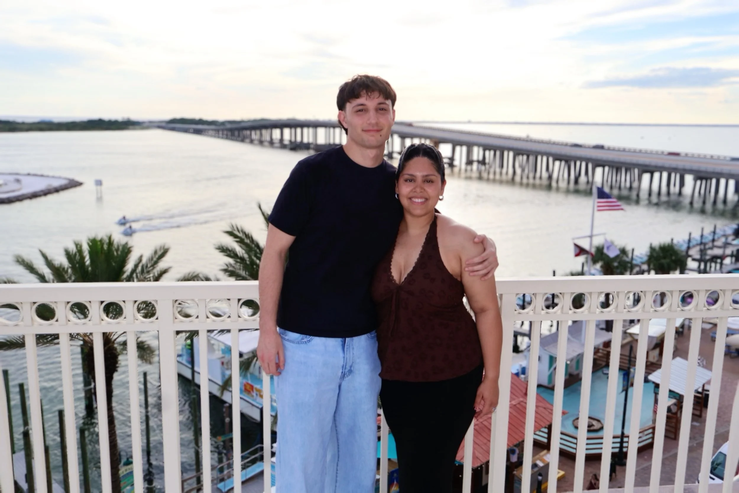 Two people, a man and a woman, standing on a balcony near water, with a bridge visible in the background during daytime.