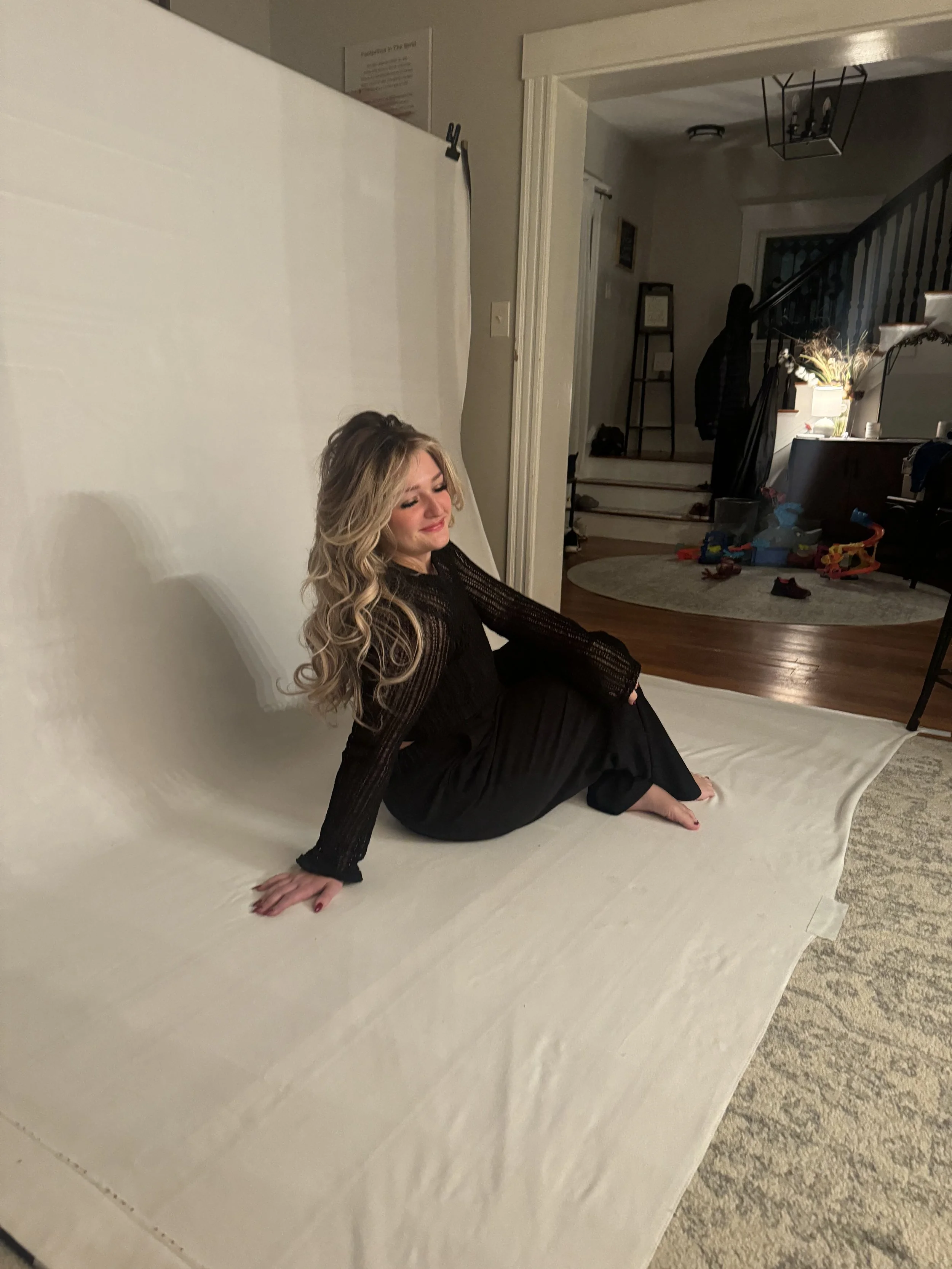 A woman with long blonde hair sitting on a white backdrop in a room, smiling with her eyes closed, dressed in a black sheer top and black pants.