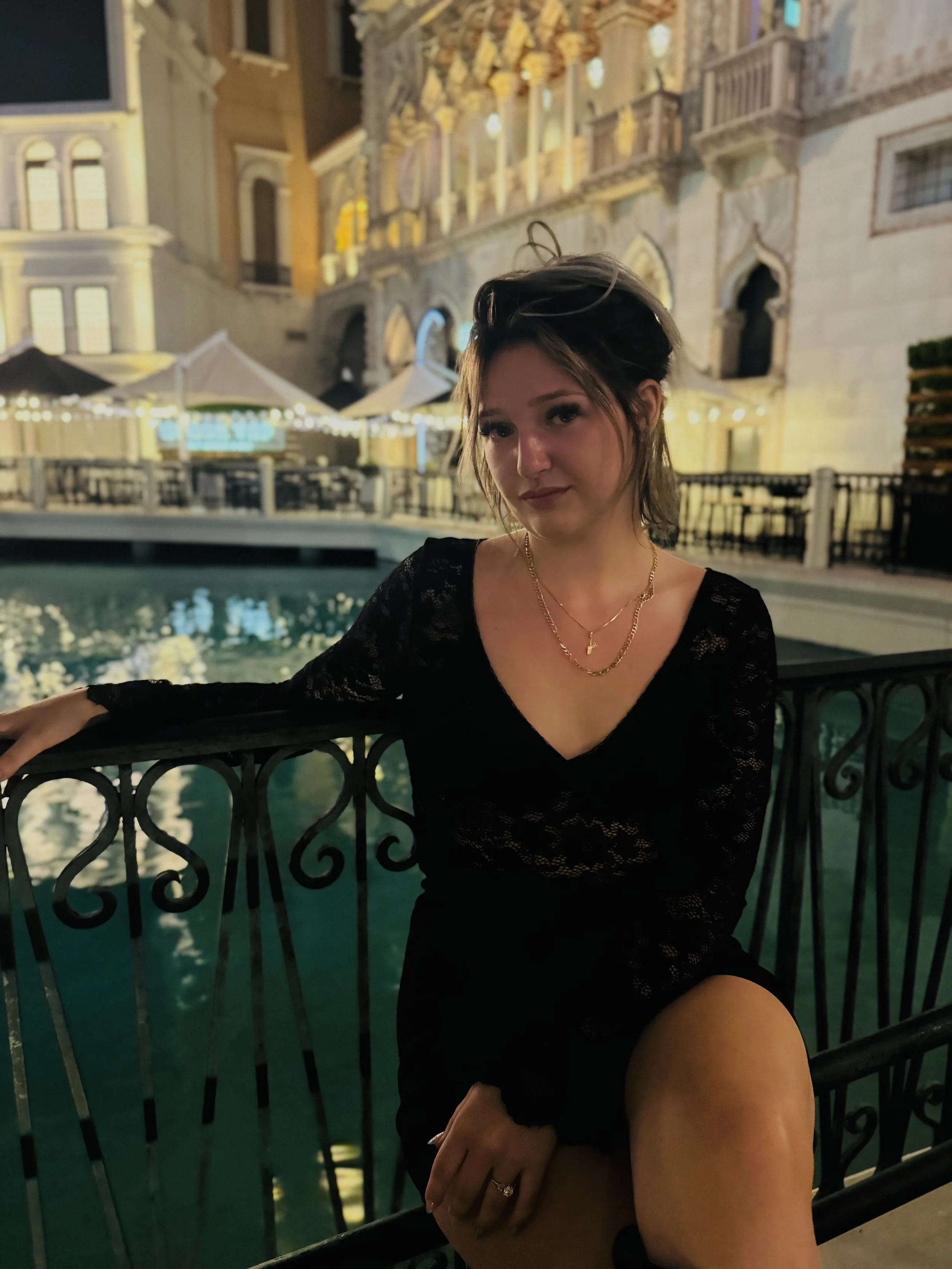 A woman in a black lace dress sitting by a decorative black wrought iron railing near a water feature at night, with illuminated historic buildings and outdoor seating with umbrellas in the background.