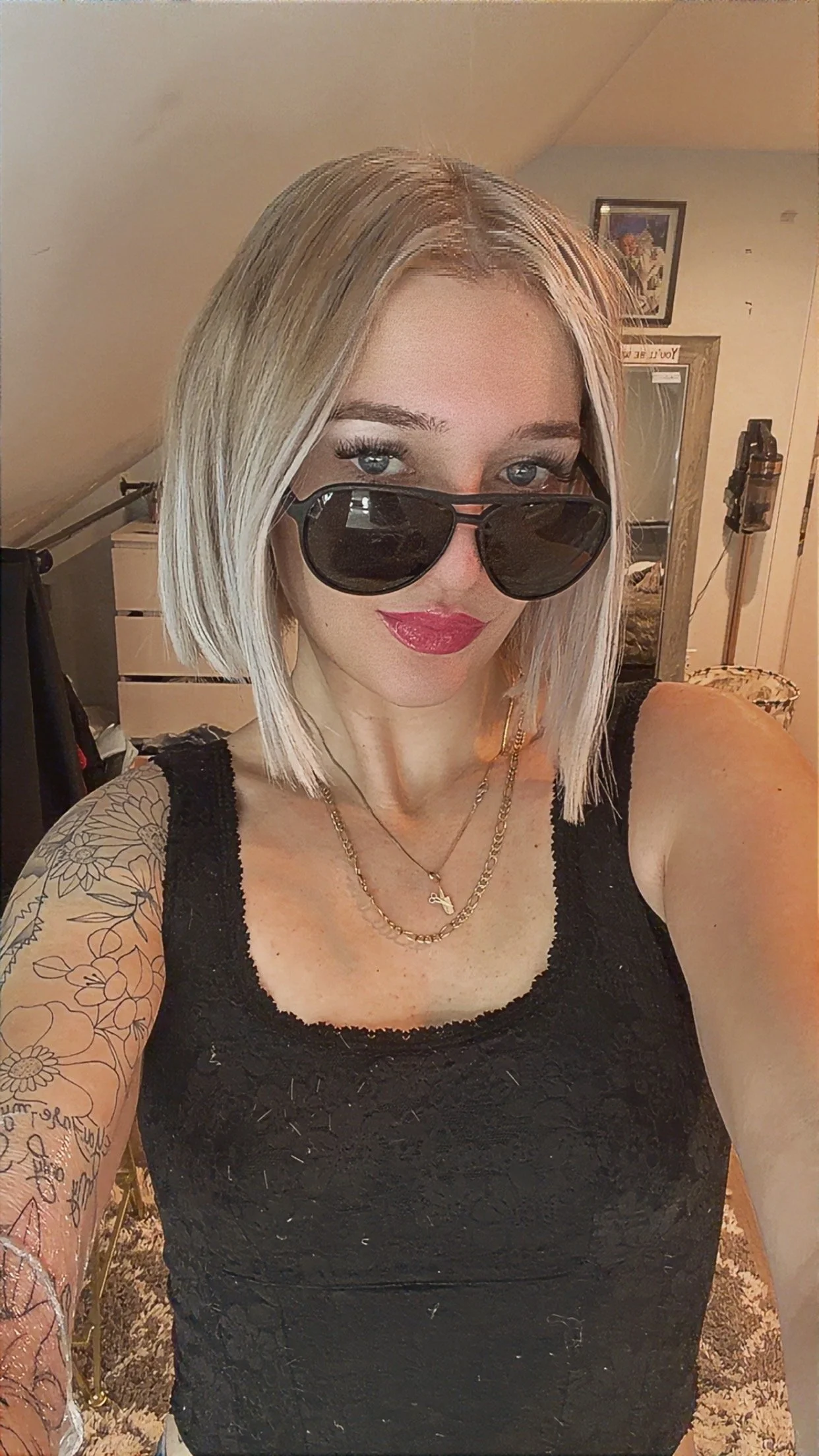 A woman with blonde, shoulder-length hair wearing aviator sunglasses, a black lace sleeveless top, and layered gold necklaces, taking a selfie in a room with a mirror, framed pictures, and furniture in the background.