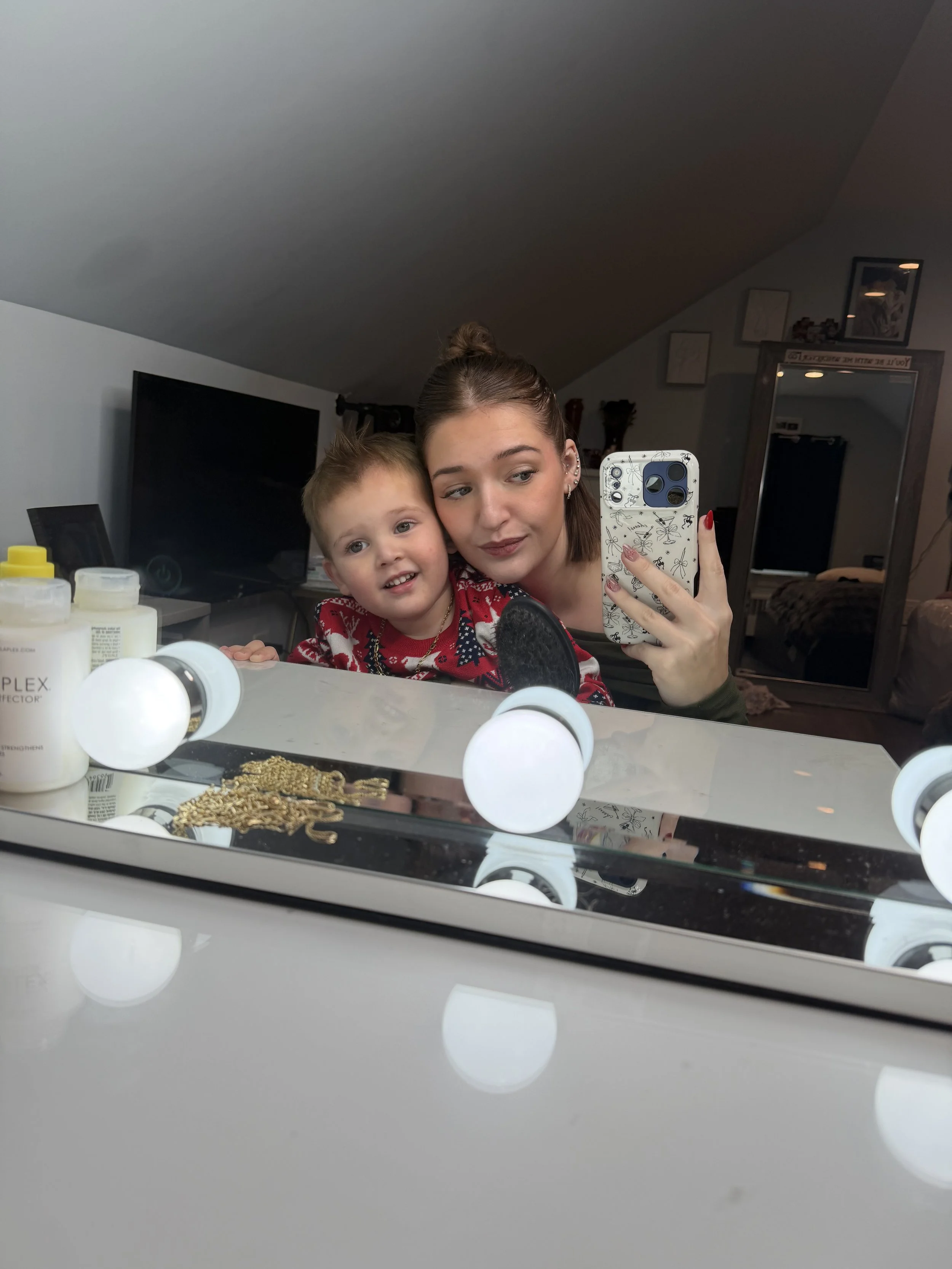 A woman and young boy taking a selfie in front of a mirror with vanity lights, in a bedroom or dressing room.