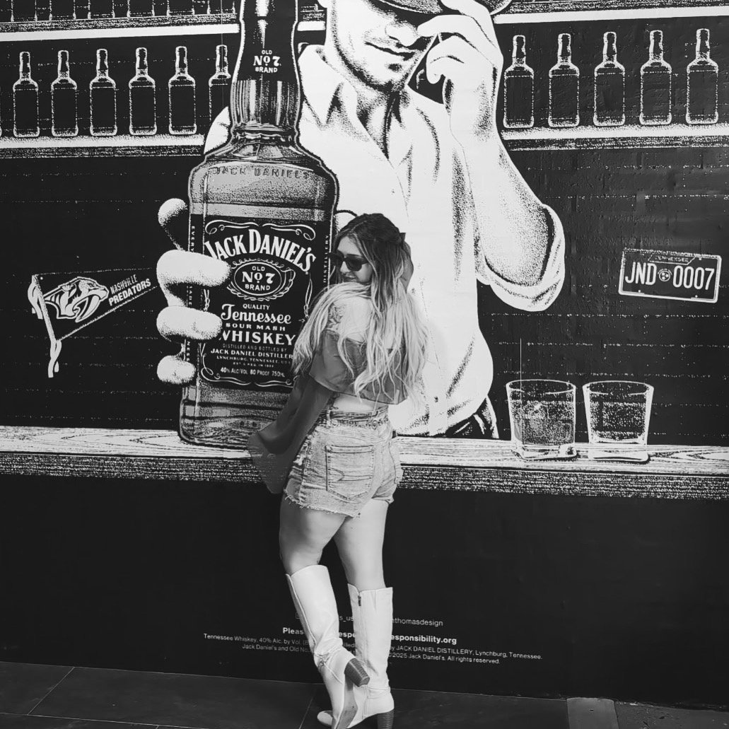 A woman with long hair, wearing sunglasses, a jacket, denim shorts, and cowboy boots, stands with her back to the camera in front of a large wall mural featuring a person holding a bottle of Jack Daniel's whiskey and Nashville Predators hockey team logo.