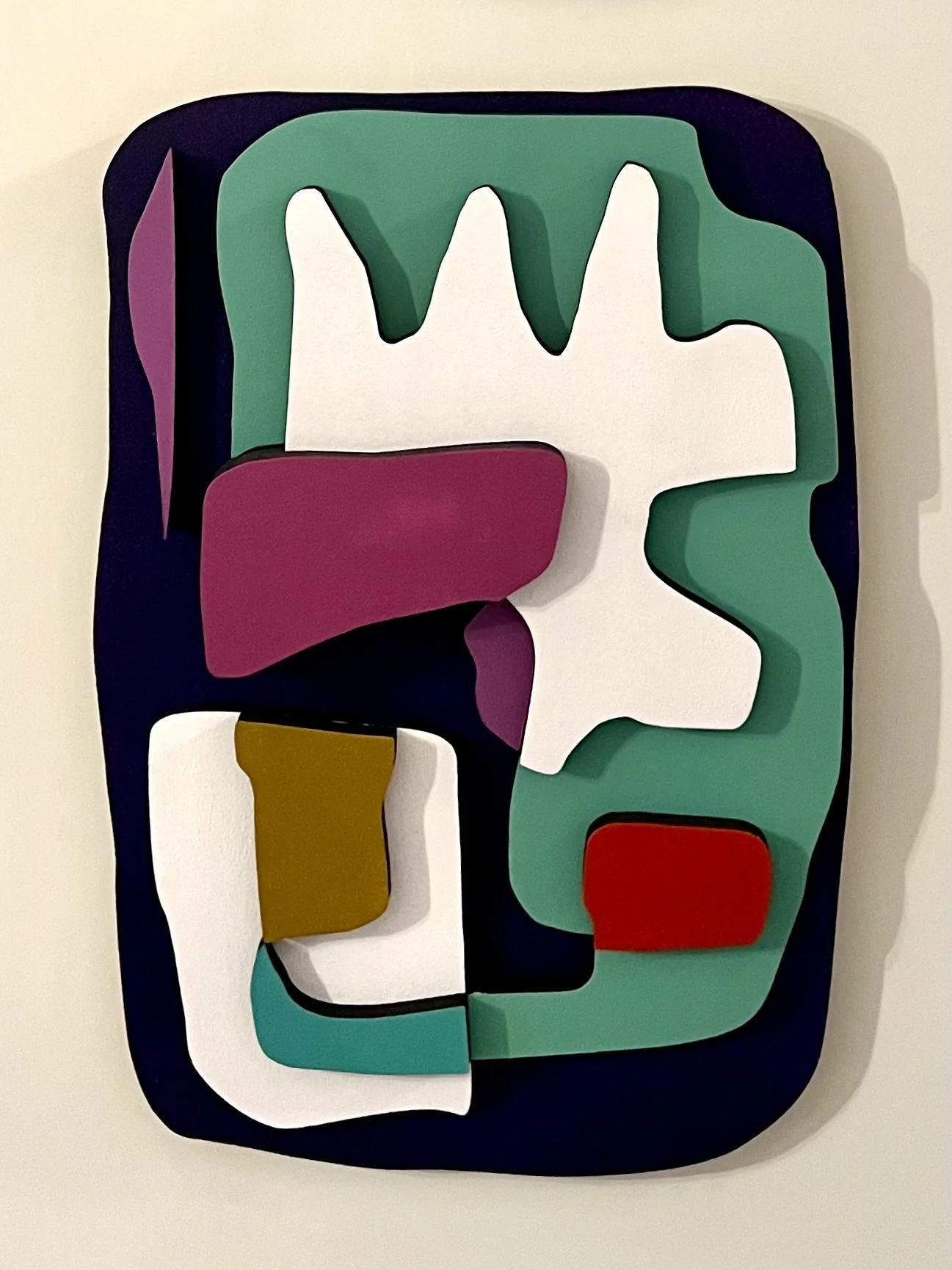 Colorful abstract mixed media art piece with various geometric and organic shapes layered with vibrant colors such as purple, green, white, red, black, brown, and teal, mounted on a rectangular black background.