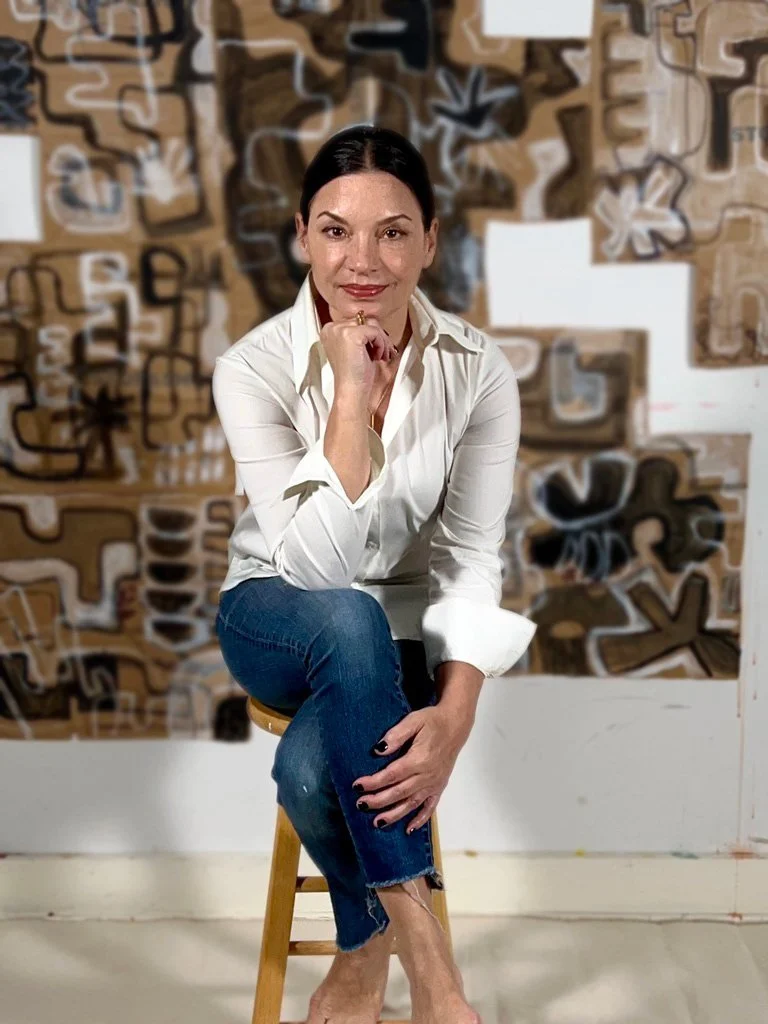 A woman with dark hair in a ponytail, wearing a white button-up shirt and blue jeans, sitting on a wooden stool in front of an abstract graffiti-style mural wall.