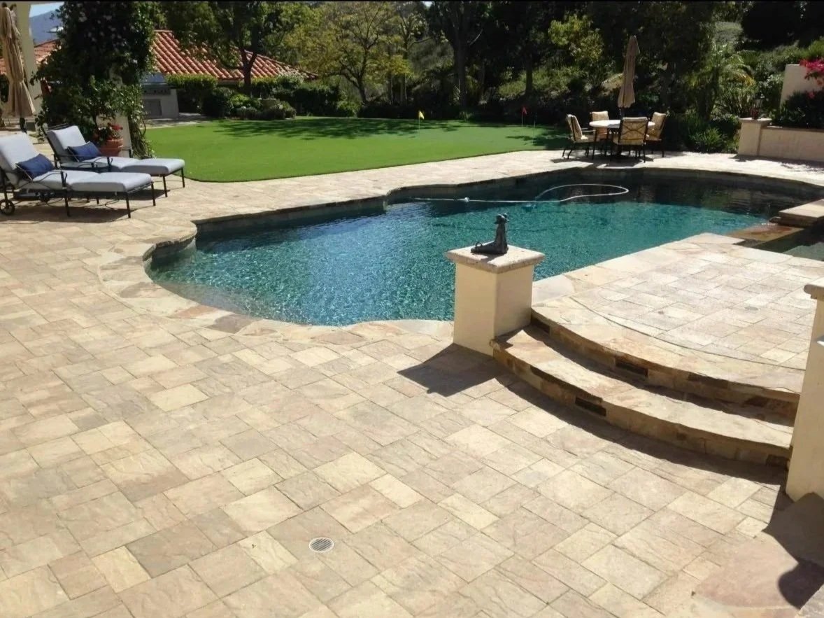 A backyard with a swimming pool, stone patio, and outdoor furniture including lounge chairs and a table with chairs, surrounded by trees and greenery.